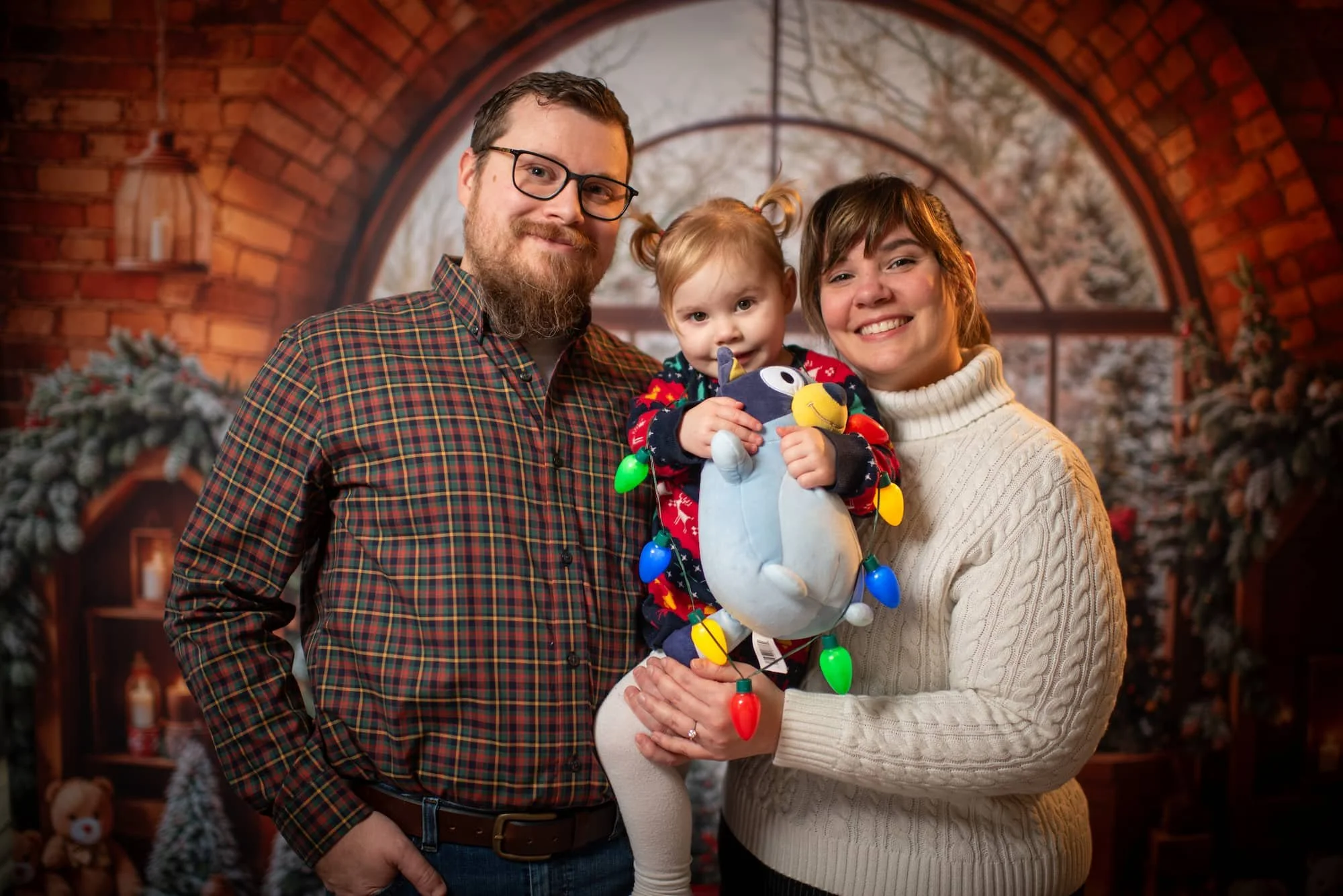 Family of three celebrating Christmas indoors with a decorated background that includes Christmas trees and ornaments. The father has glasses, a beard, and is wearing a plaid shirt. The mother has short hair and a sweater. The young girl, dressed in a festive sweater, is holding a large penguin stuffed toy decorated with colorful Christmas lights.