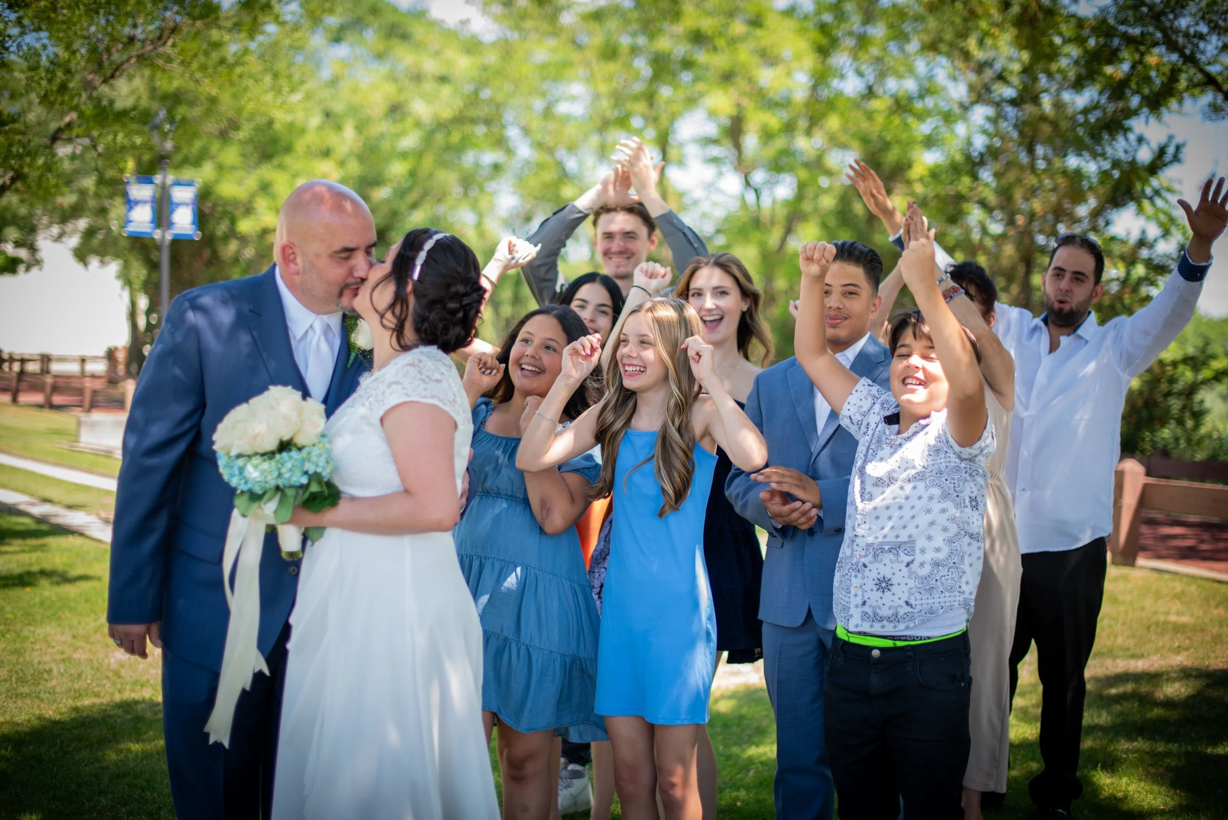 A bride and groom share a kiss outdoors with wedding guests celebrating around them on a sunny day.