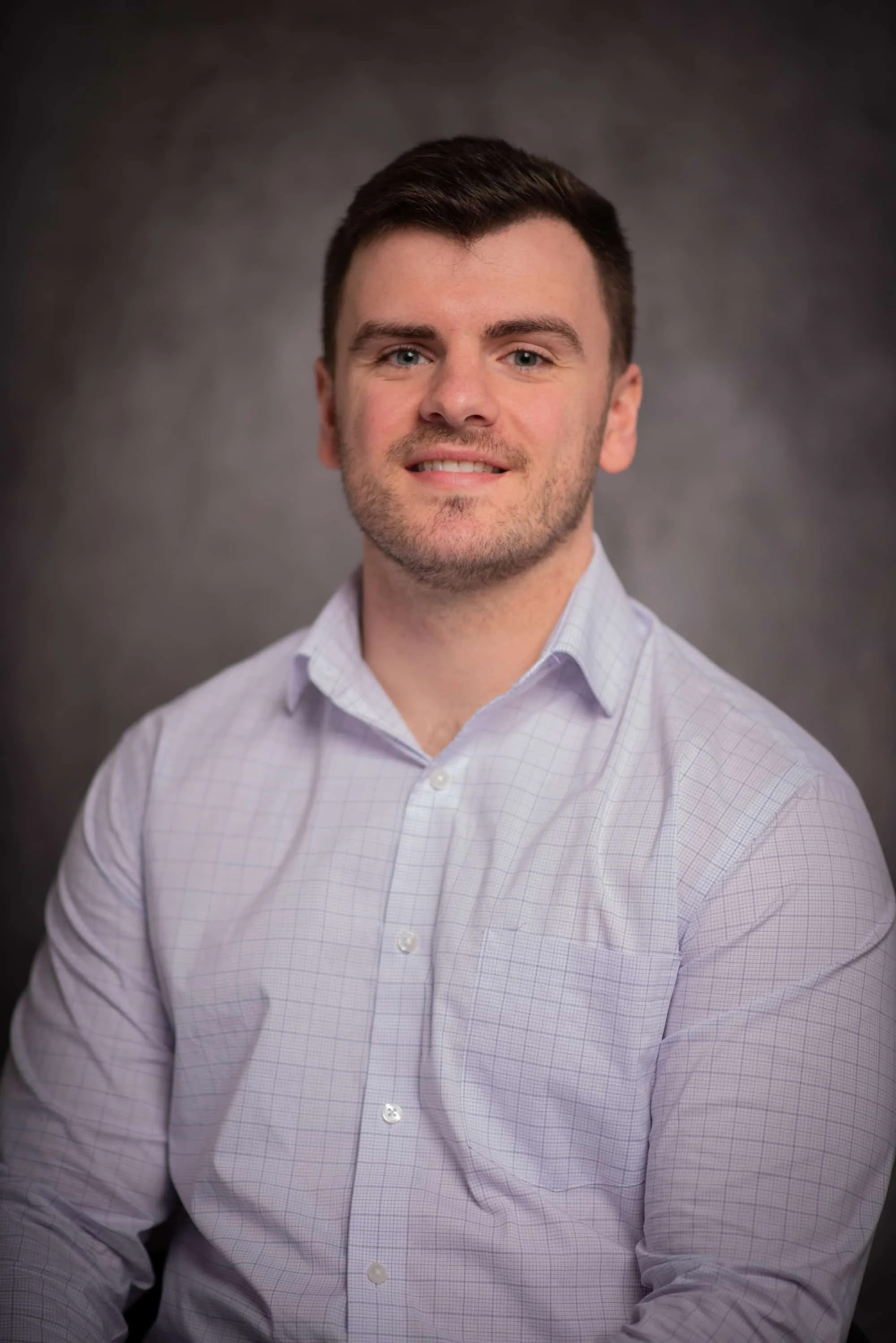 Portrait of a young man with dark hair, wearing a light-colored dress shirt, smiling, against a dark gray background.