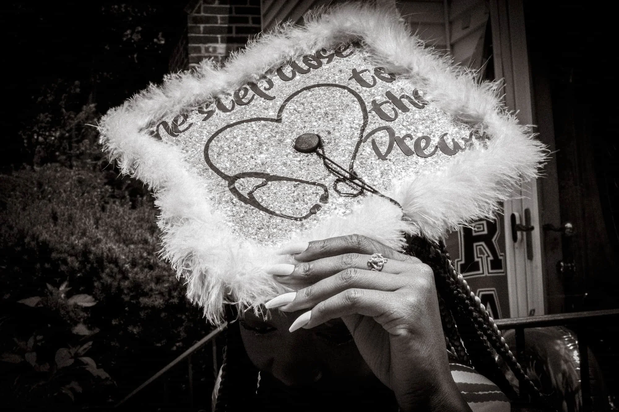 Person holding a decorated graduation cap with a heart and text, surrounded by fluffy trim, in black and white.