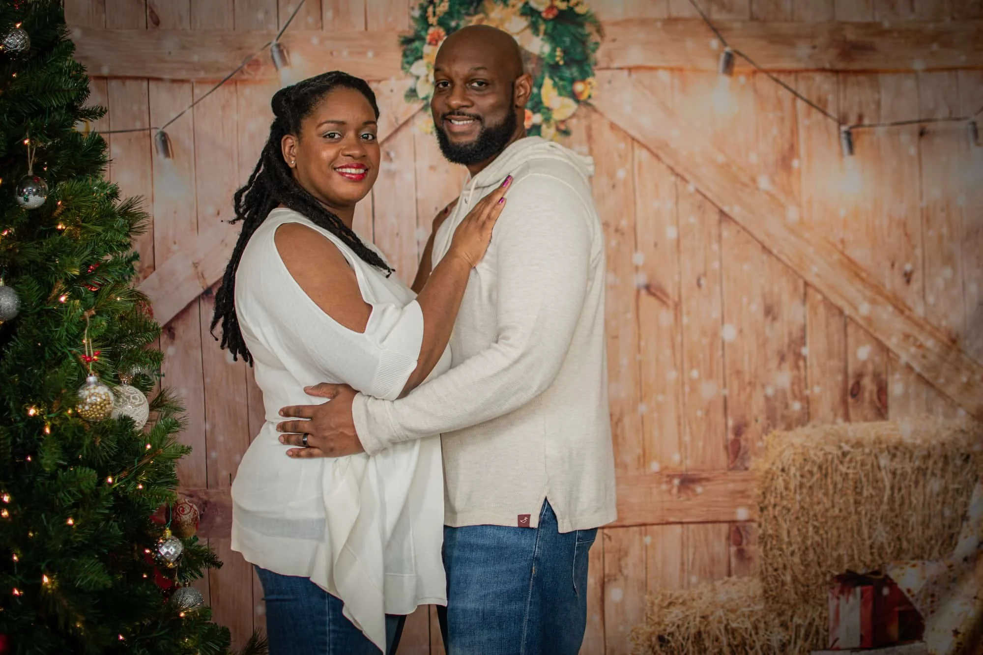 A couple standing close together, smiling, with Christmas decorations including a decorated tree, wrapped gifts, and a wooden wall background, suggesting a holiday setting.