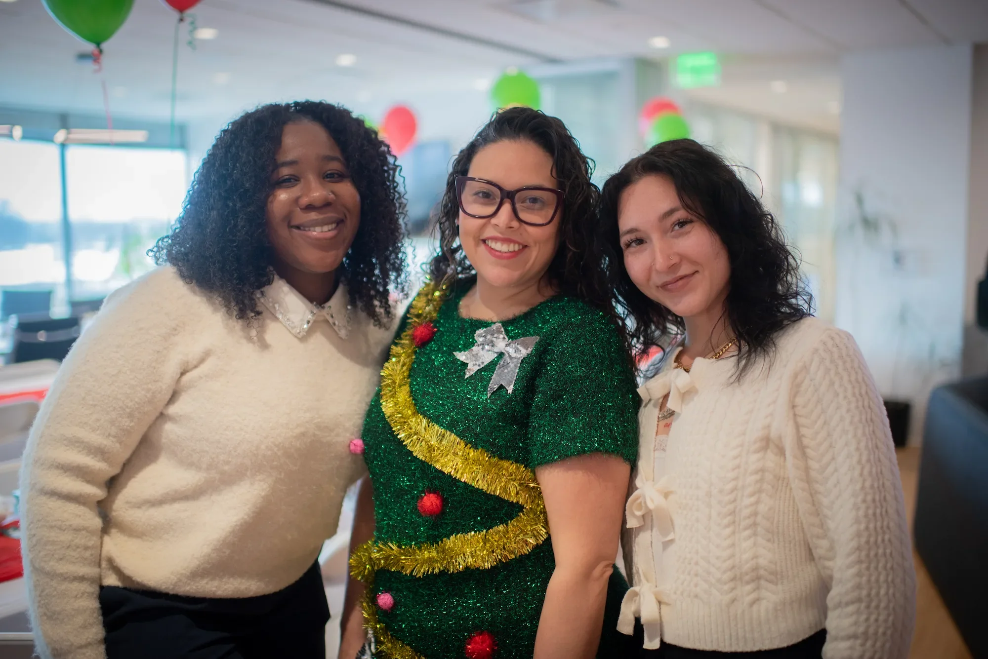 Three women smiling, celebrating Christmas indoors, with balloons and decorations in the background. One wears a Christmas tree costume adorned with tinsel and ornaments, while the other two wear cozy sweaters.