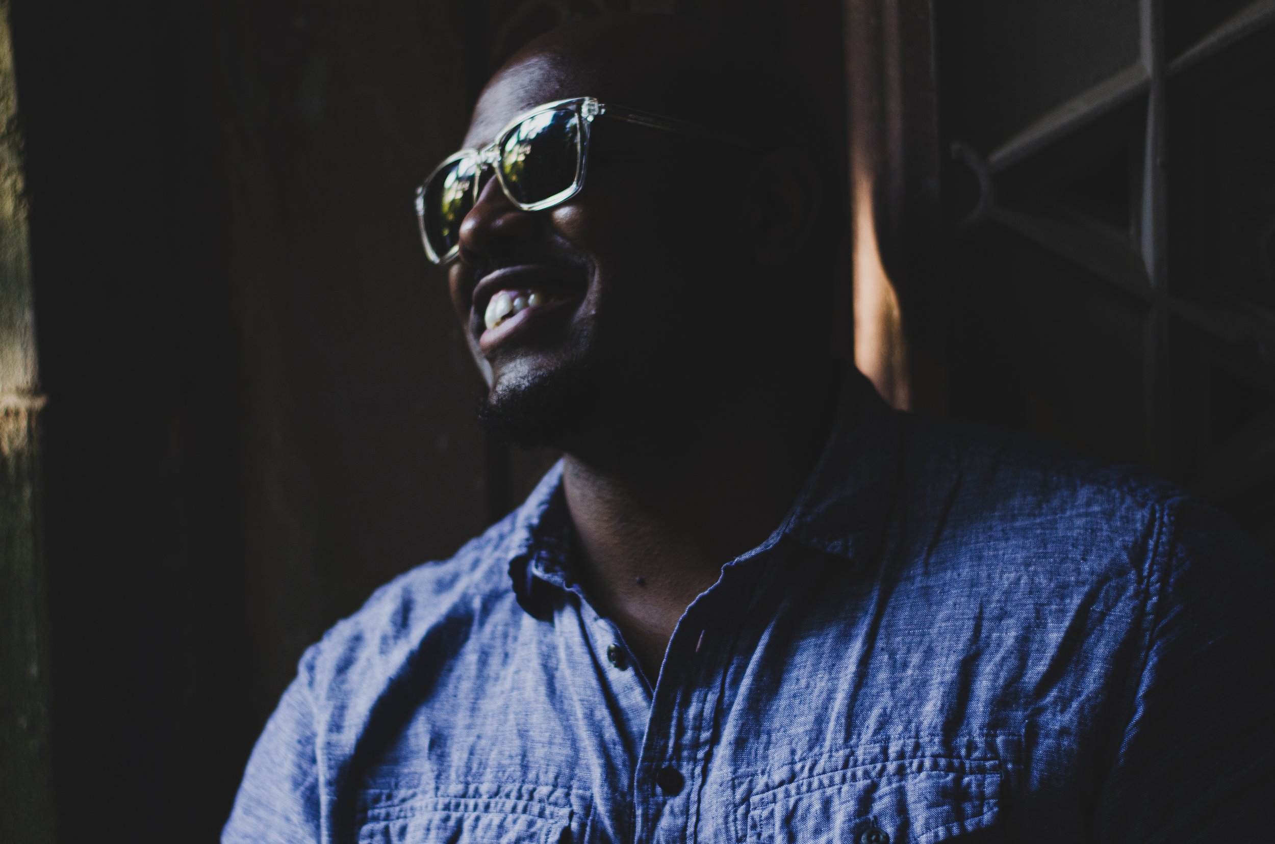A man wearing sunglasses and a blue denim shirt smiling in a shadowed setting with a dark background.
