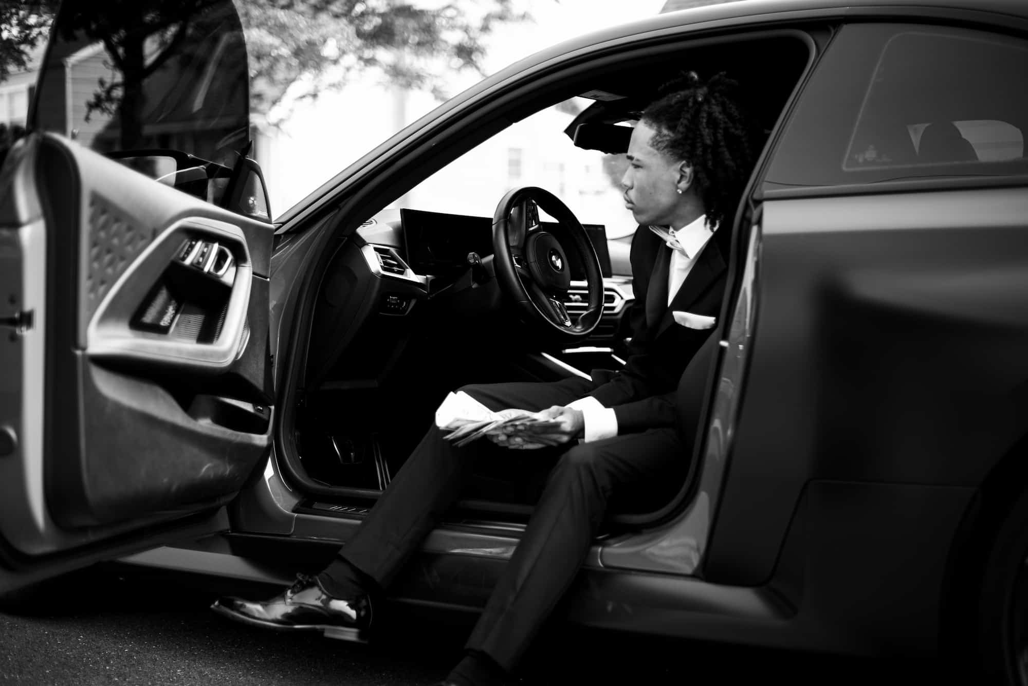 A man in a suit sitting inside a luxury car, holding a stack of papers, with the car's door open.