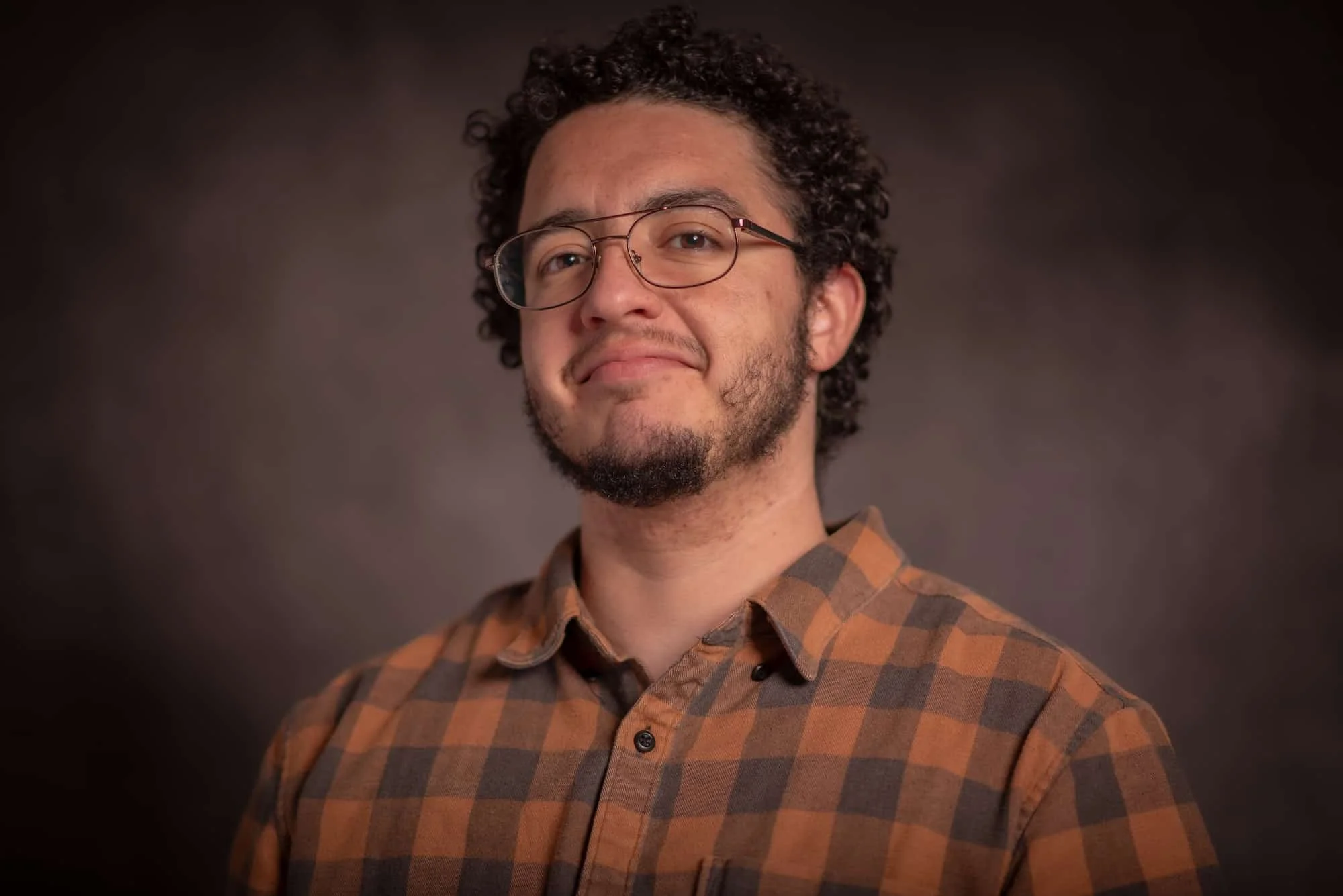 A man with curly hair, glasses, and a beard wearing a brown and orange plaid shirt against a dark background.