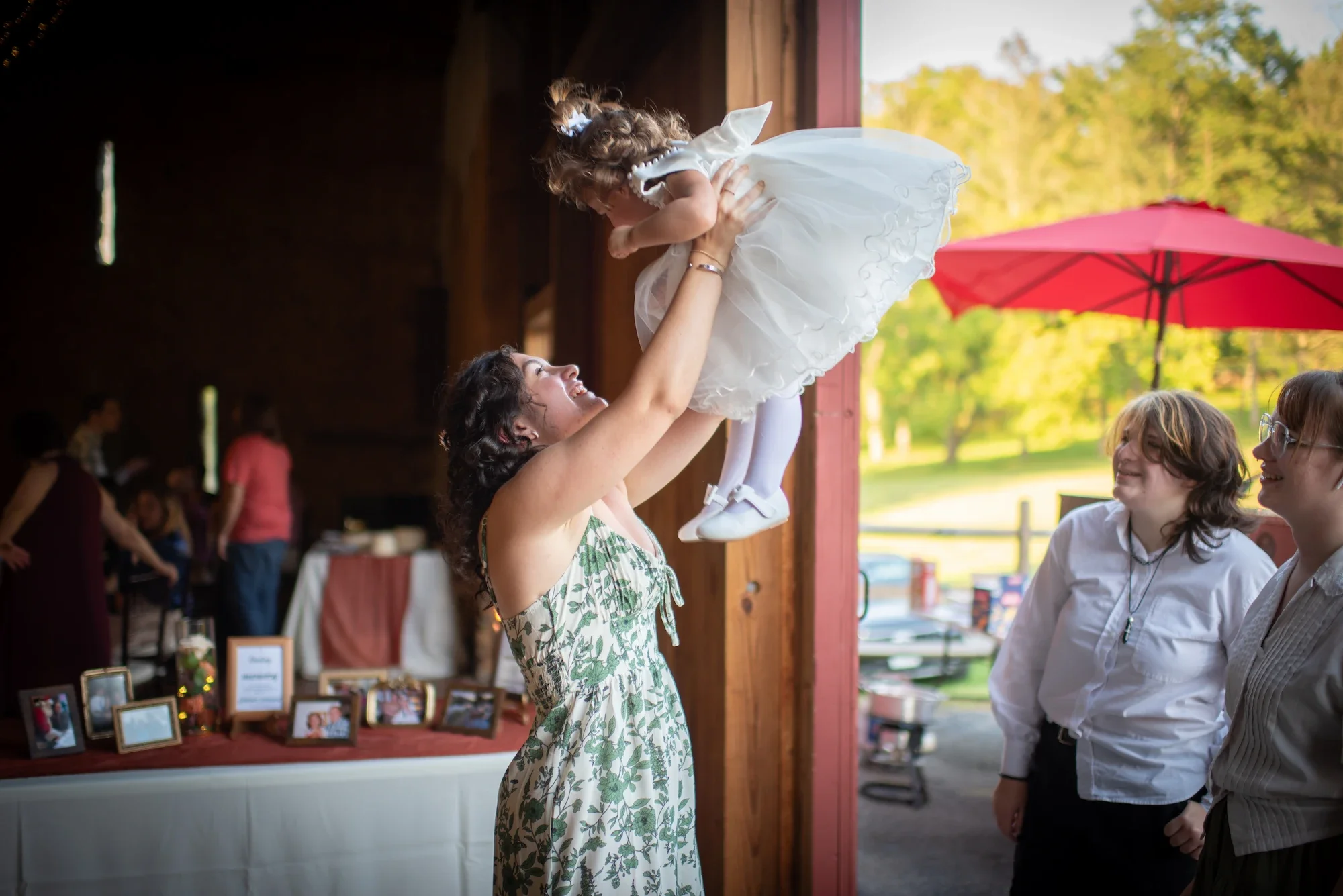 A woman lifts a young girl in a white dress with her arms stretched out, inside a rustic barn with open doors, while two women look on outside in a sunny farm setting.
