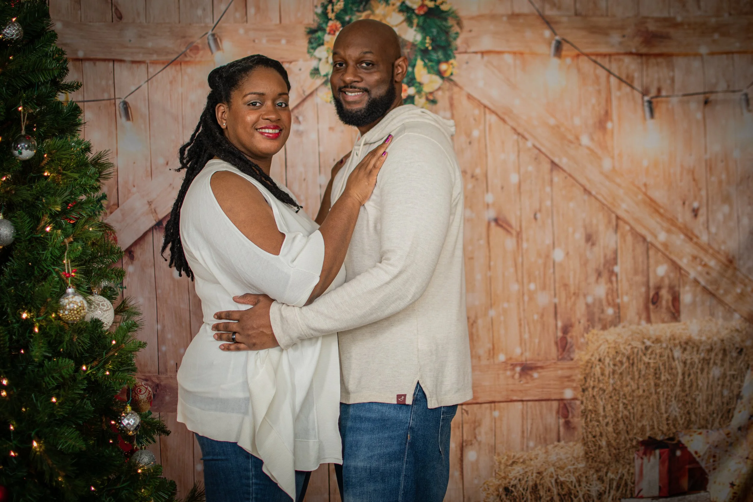 A couple standing close together indoors with a Christmas tree decorated with ornaments on the left and rustic wooden panels in the background.