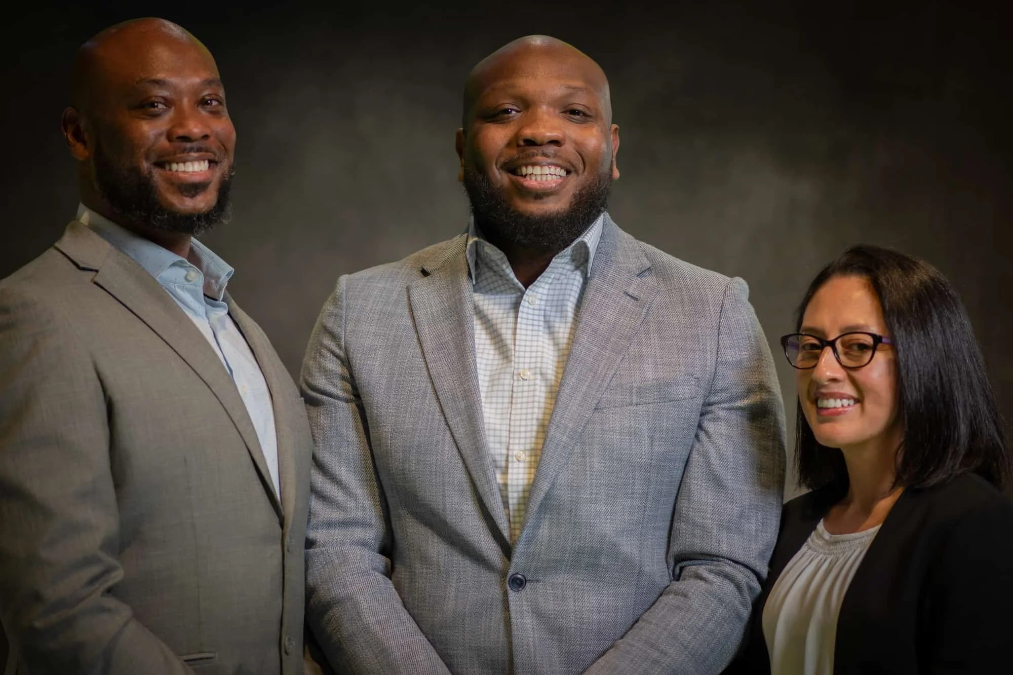 Three professionals in business attire smiling for a photo against a dark background.
