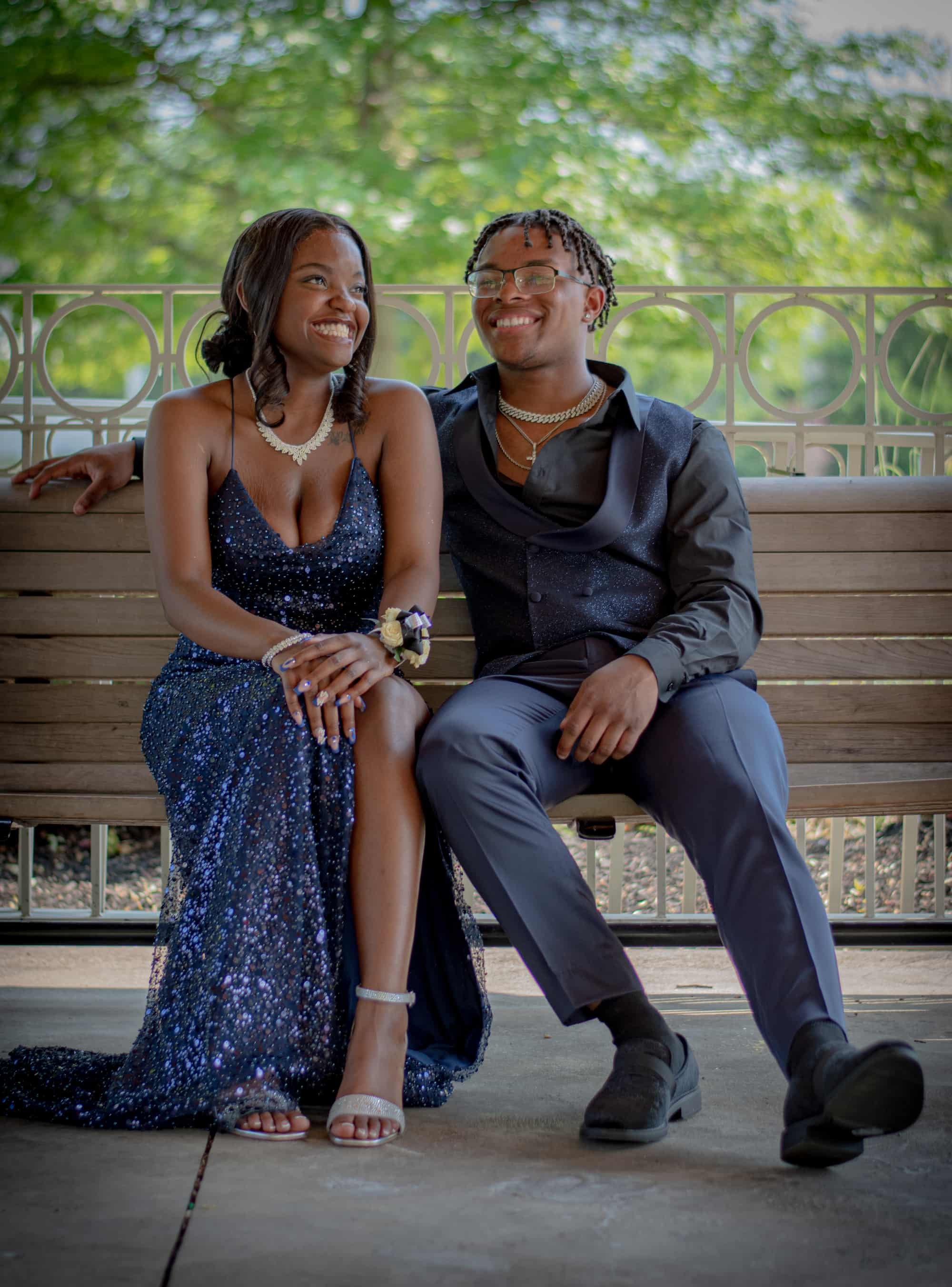 A young couple dressed formally sitting on a wooden park bench, smiling and looking at each other with a green, leafy background.
