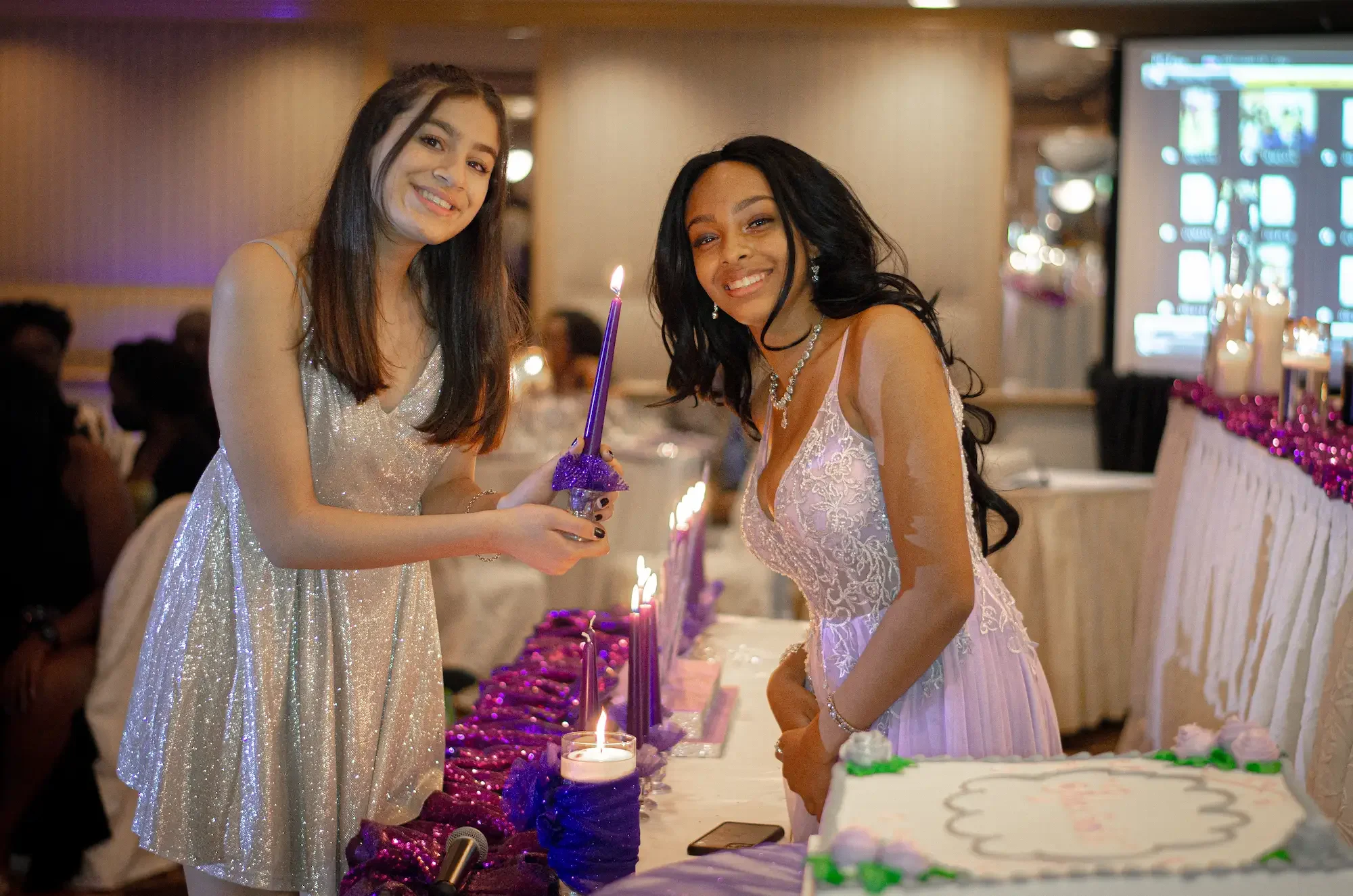 Two women in dresses at a celebration, one is lighting a purple candle, with a large decorated cake and purple and pink decorations on a table at a party or reception.