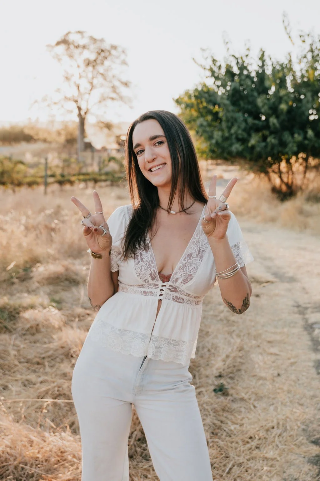 Young woman smiling and making peace signs with both hands outdoors during sunset, wearing a white lace top and white pants.