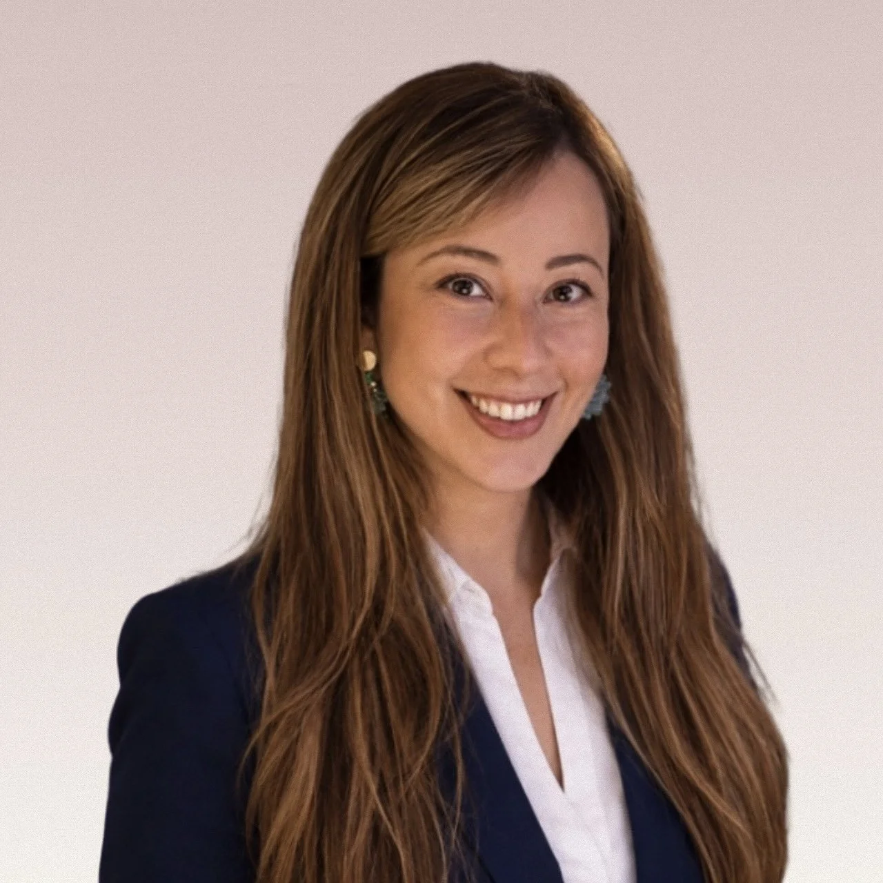 A professional woman with long reddish-brown hair, smiling, wearing a navy blazer, white blouse, and earrings, against a plain light background.