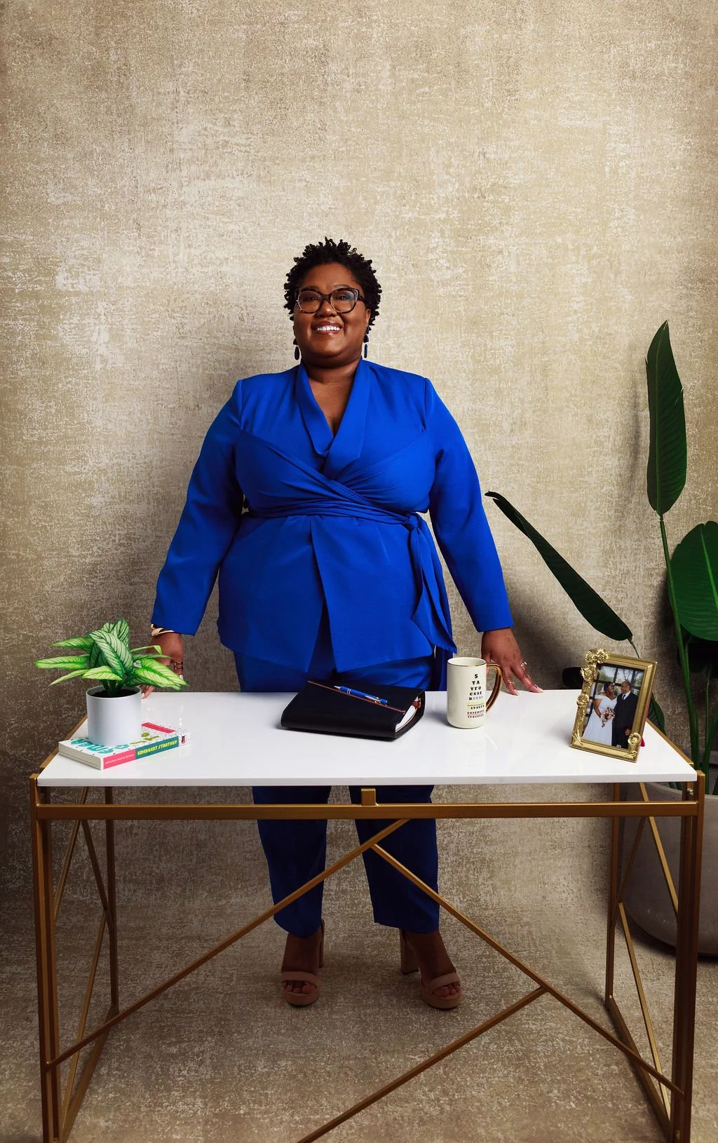 A woman dressed in a royal blue suit standing behind a white desk with a potted plant, a black notebook, a white mug, and a framed photograph, in front of a beige textured wall