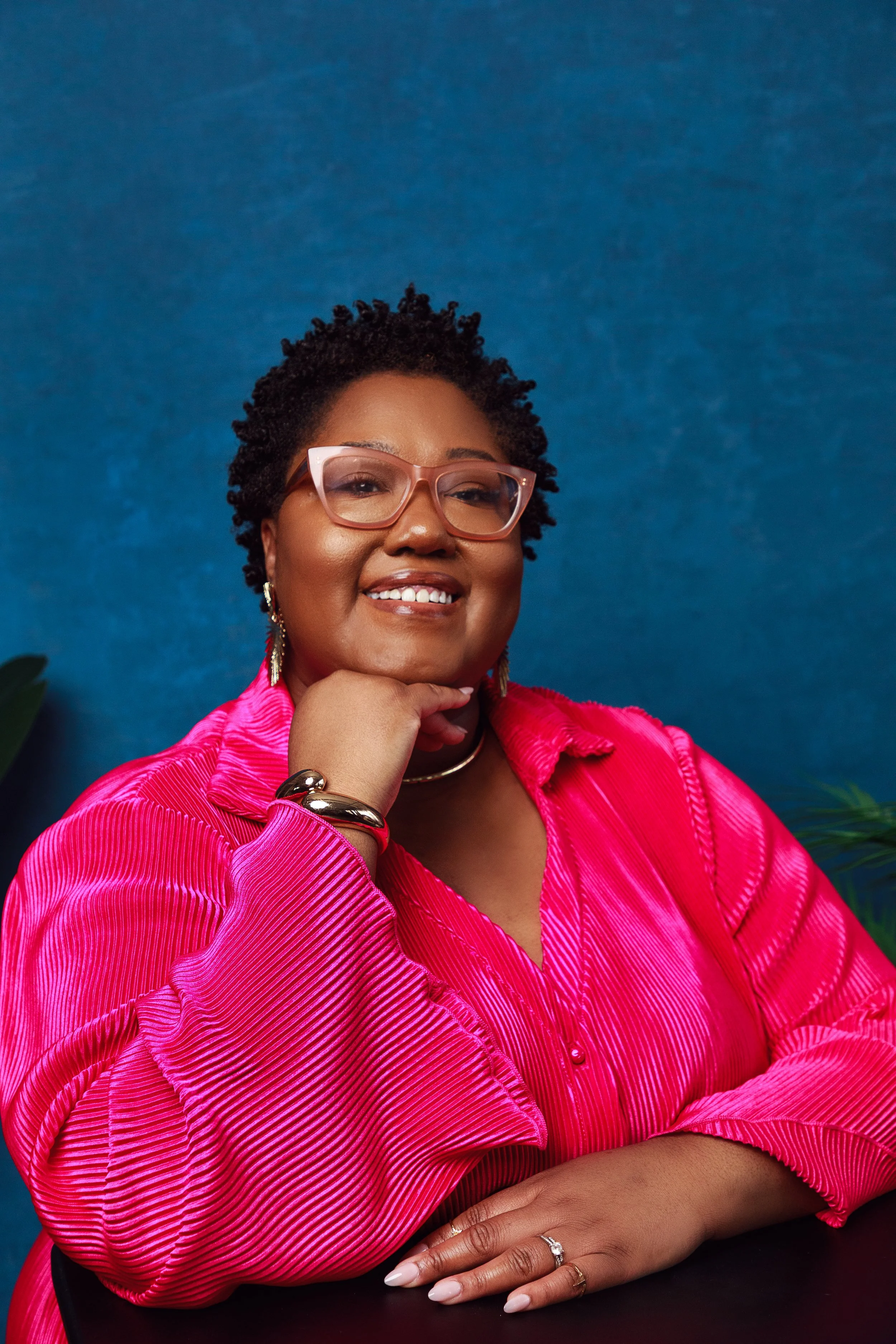 A confident woman with short curly hair wearing glasses and a vibrant pink blouse, smiling and resting her chin on her hand, posed against a blue background.