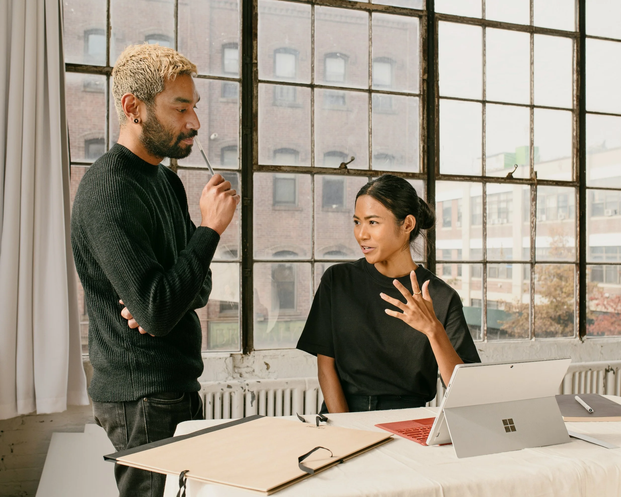 A man and woman in an office engaged in conversation. The woman appears to be explaining something, gesturing with her hand, while the man listens thoughtfully, holding a pen near his face. They are using a tablet and have documents and a pen on the table, with a large industrial-style window behind them showing a cityscape.