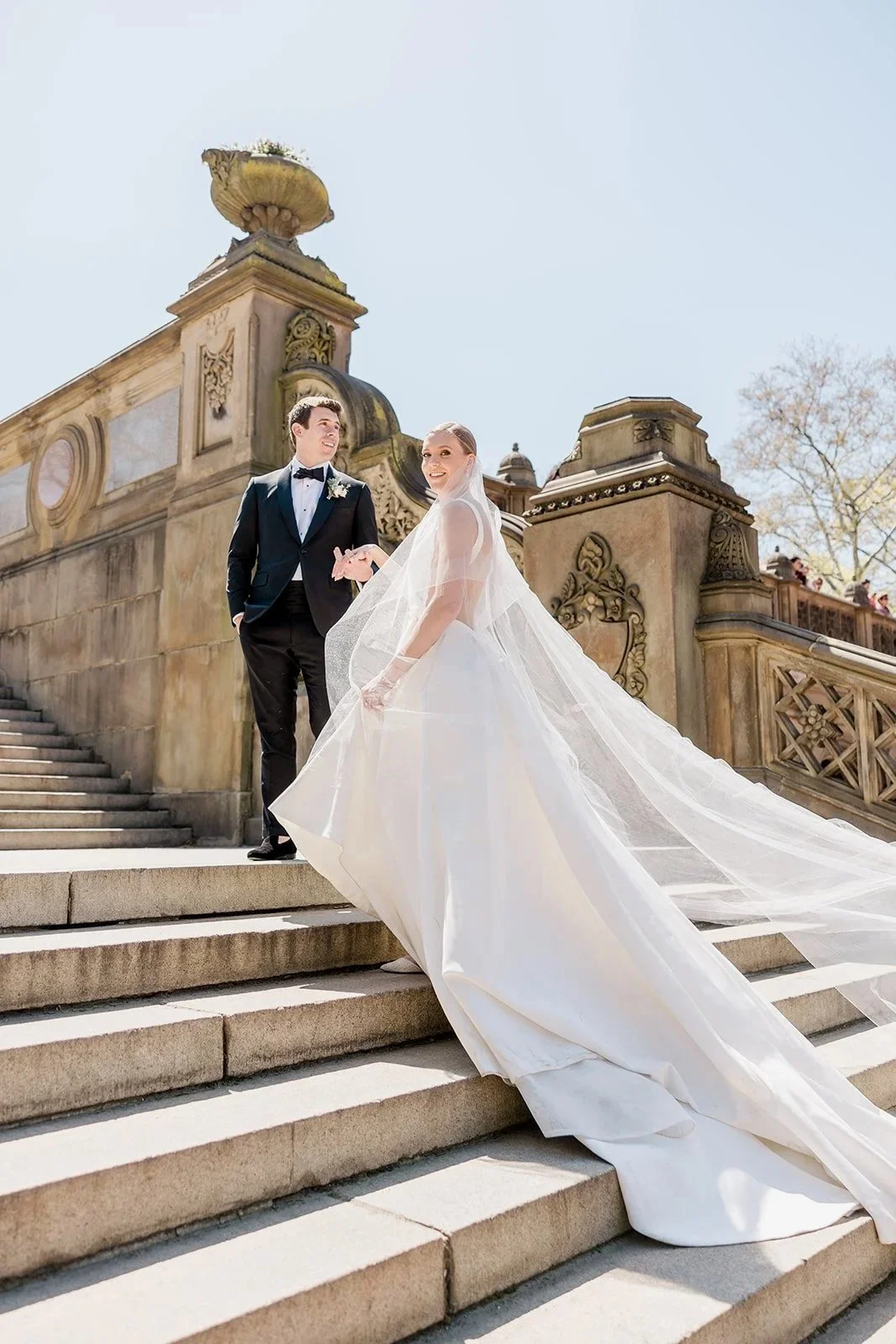 Bride and groom standing on outdoor stone staircase, with bride in a long white wedding gown and veil, groom in a black tuxedo with bow tie, historic building with ornate architecture in background on a sunny day.