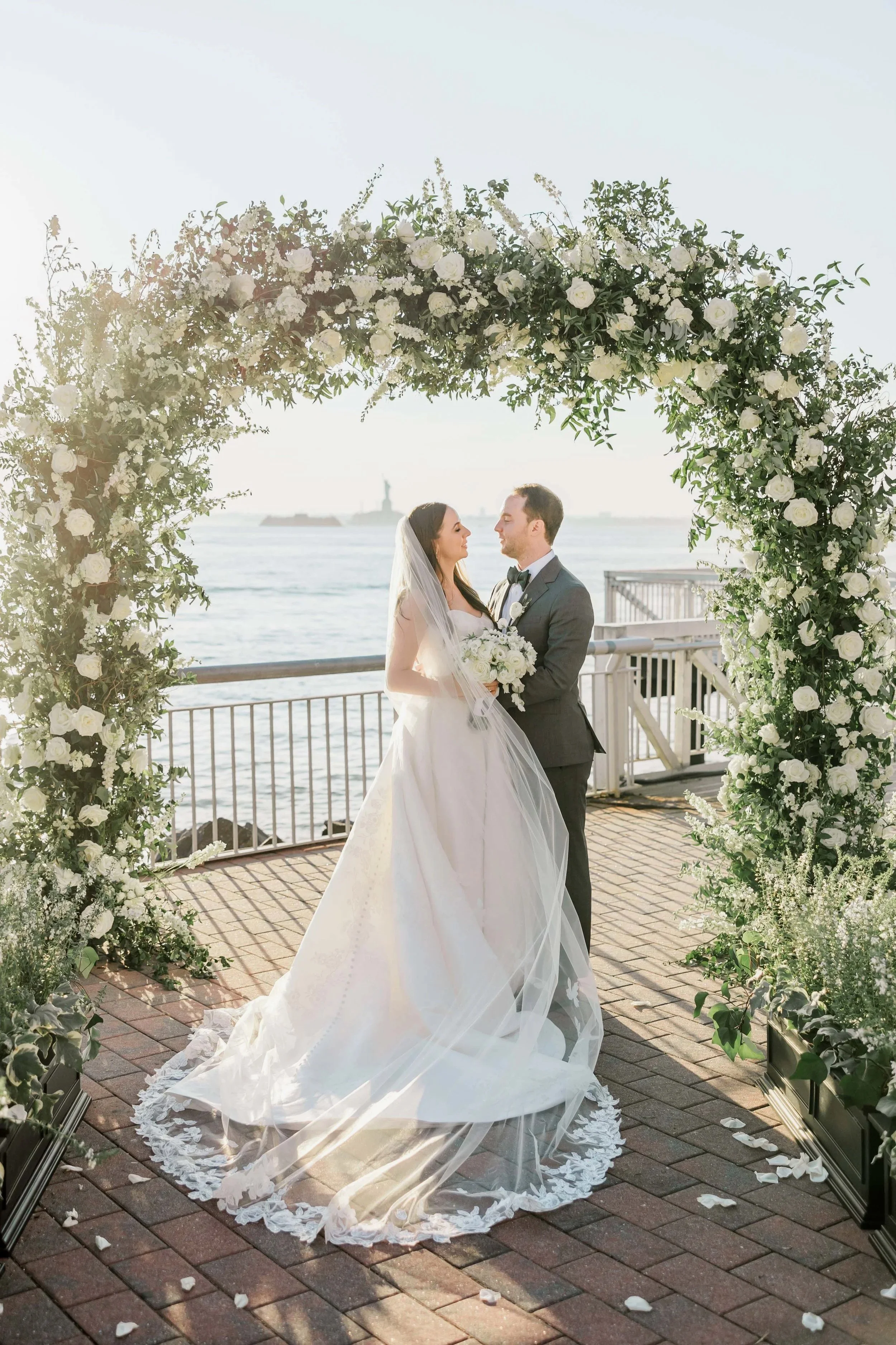 Bride and groom standing under a floral arch by the water, holding hands and gazing at each other on their wedding day.