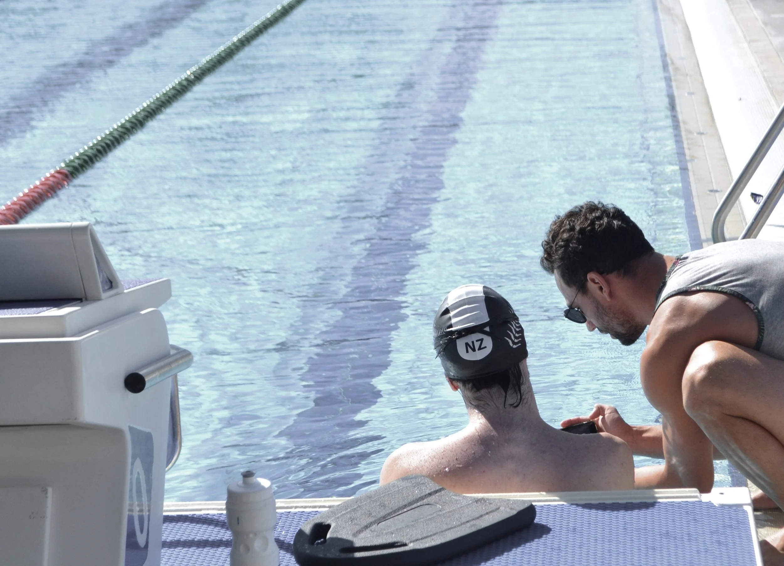 Swimmer with NZ cap sitting at the edge of an outdoor pool, talking with a man beside him.