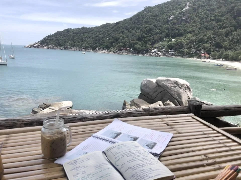 A table with an open notebook, papers, and a jar of iced coffee overlooking a bay with rocks, boats, and a green hillside.