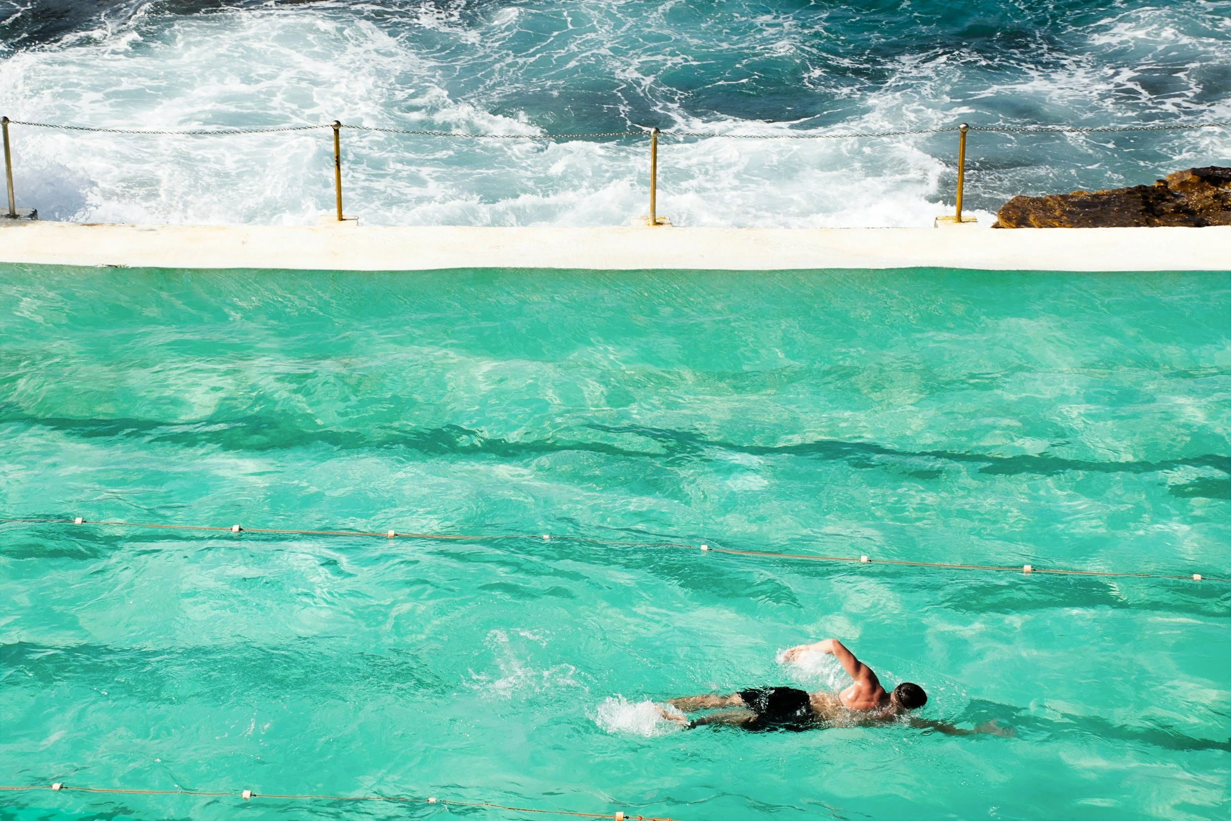 A person swimming in a turquoise outdoor pool with a white edge, located near the ocean, with waves crashing against the rocks in the background.