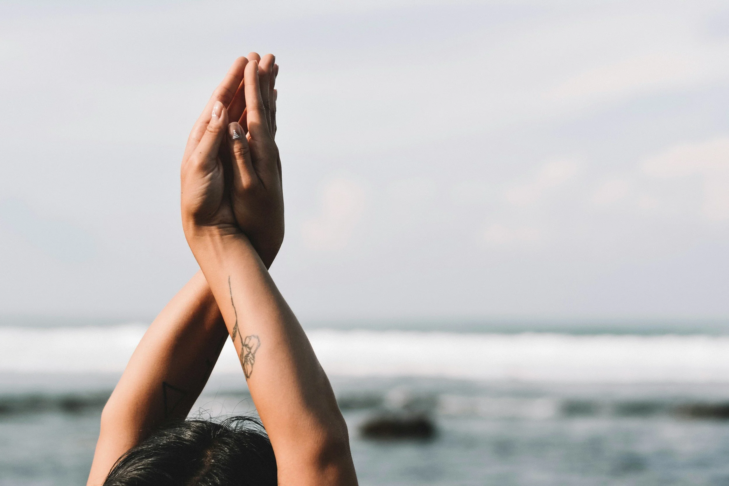 Person with tattoos doing yoga or meditation outdoors near the ocean with hands raised in prayer position.