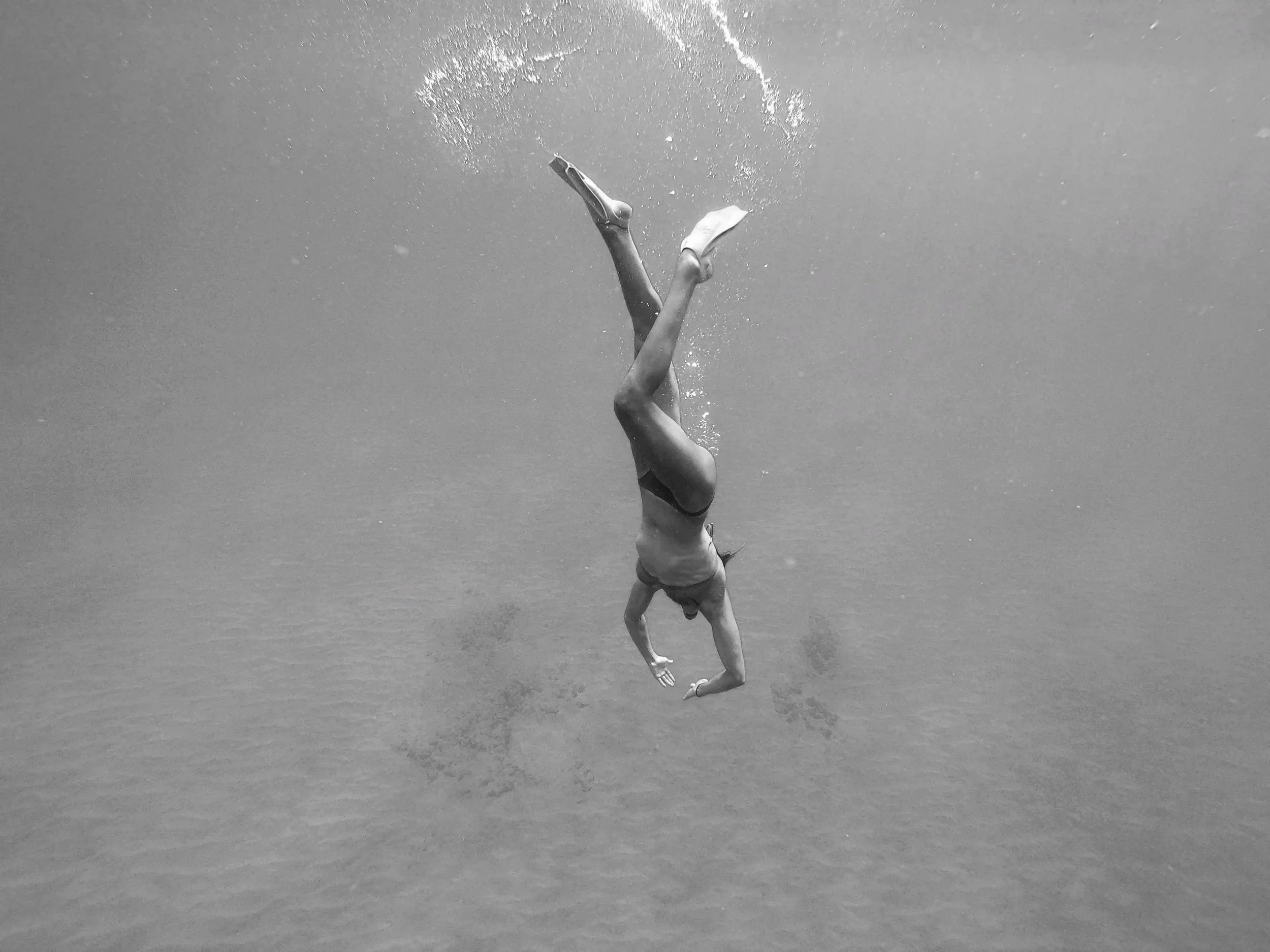 Person diving underwater, upside down with legs pointing upward and head pointing downward, wearing swimwear, surrounded by bubbles and water.