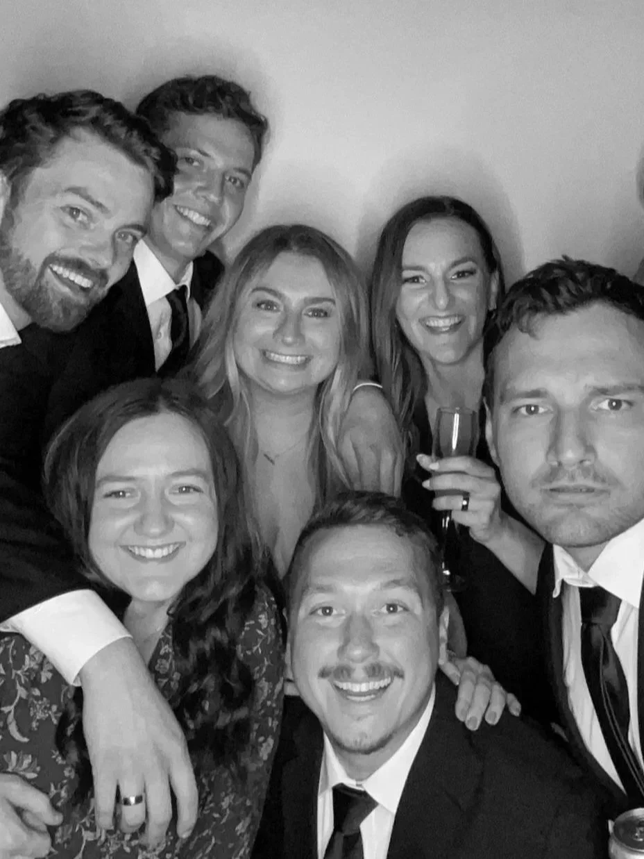 Group of people in formal attire smiling and taking a selfie at a party or gathering, with some holding champagne glasses.