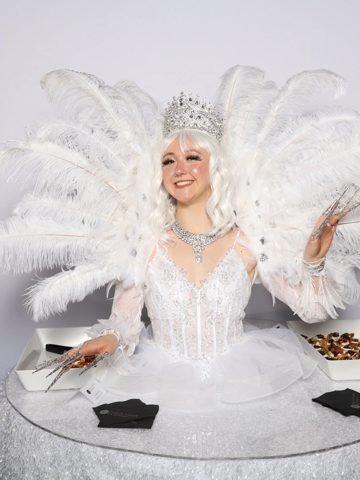 A woman dressed in a white gown with lace and beadwork, wearing a sparkling tiara and necklace, and surrounded by large white feathers, posing in a photo booth in Moorhead, Minnesota.