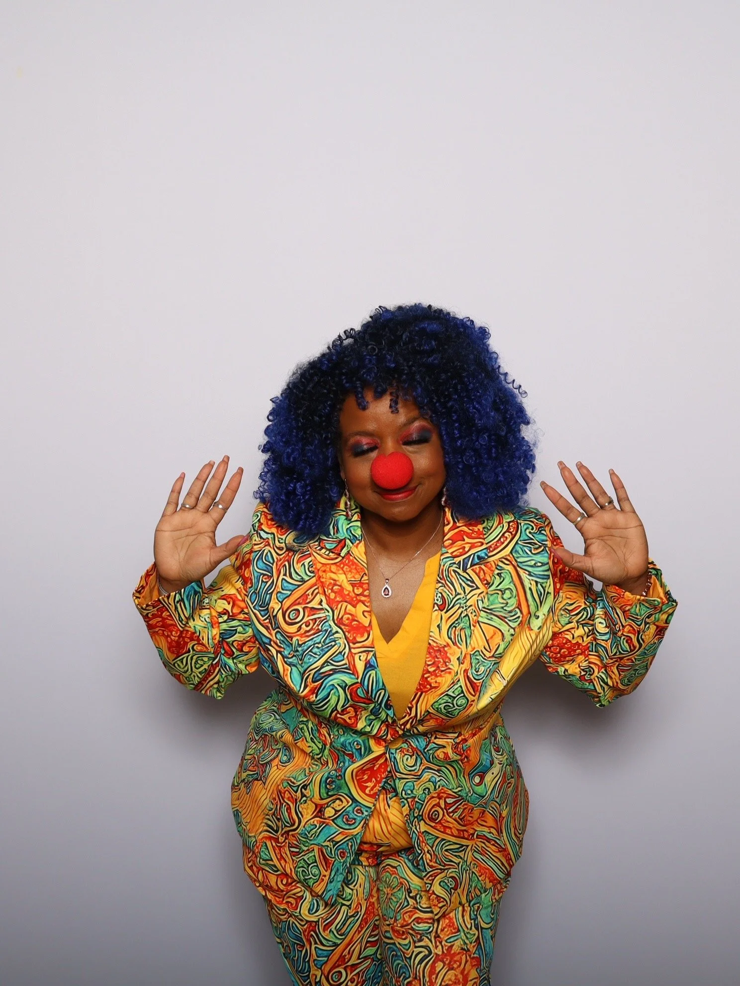 A woman dressed as a clown with curly hair, a red clown nose, colorful patterned jacket, and a yellow top, posing against a plain white wall with her hands raised. She is posing in a photo booth in Fargo, ND.
