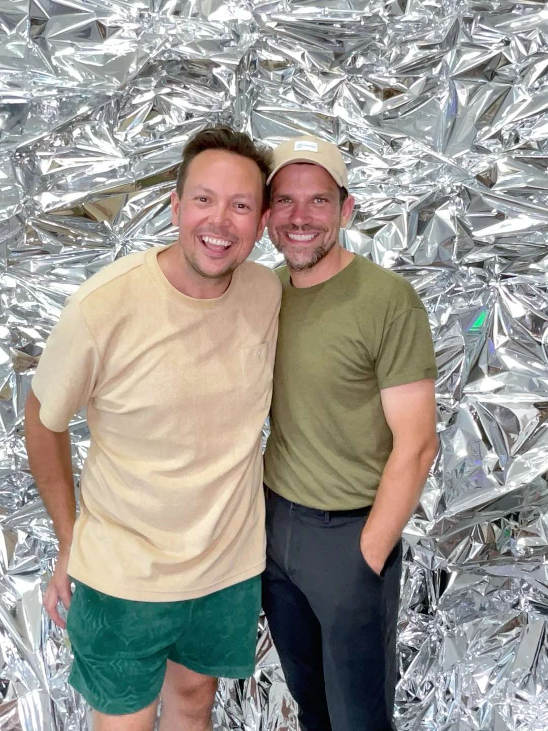 Two men smiling and standing close together in front of a shiny silver crinkled foil backdrop.