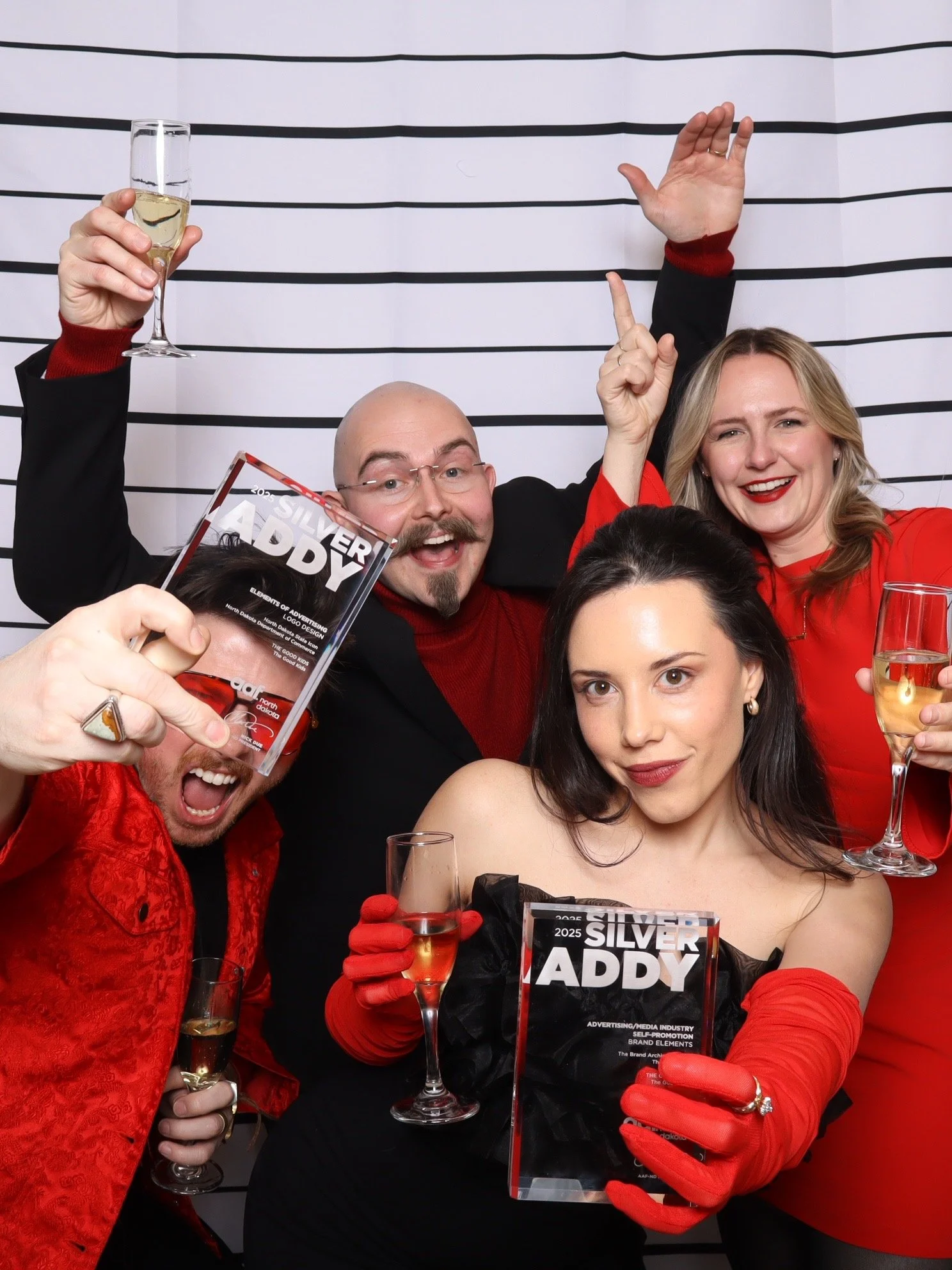 Group of people celebrating and posing in a photo booth with glasses of champagne, holding awards titled 'Silver Addy', in front of a striped black and white background.