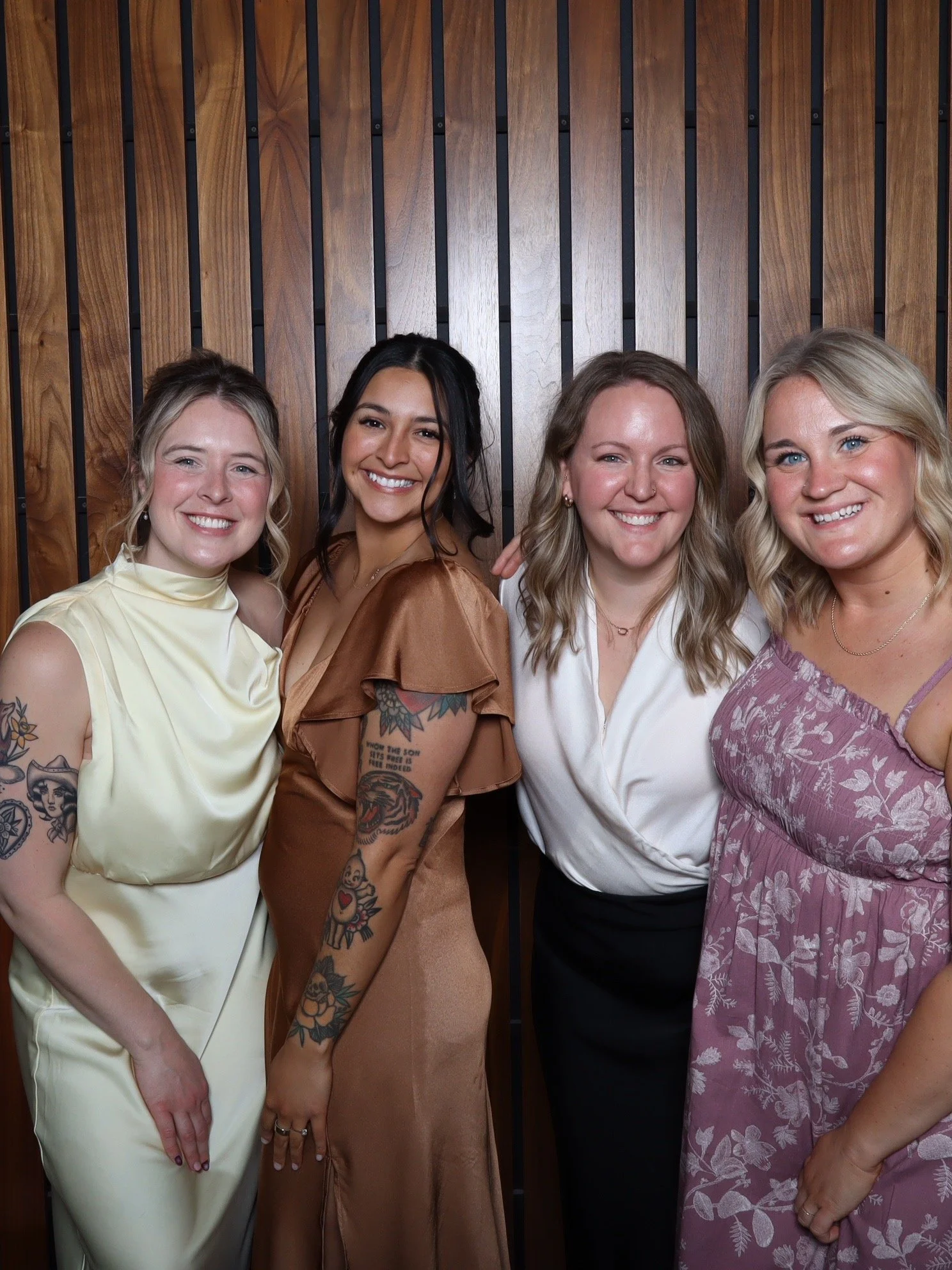 Four women standing together in front of a wooden wall, smiling at the camera of a photo booth.