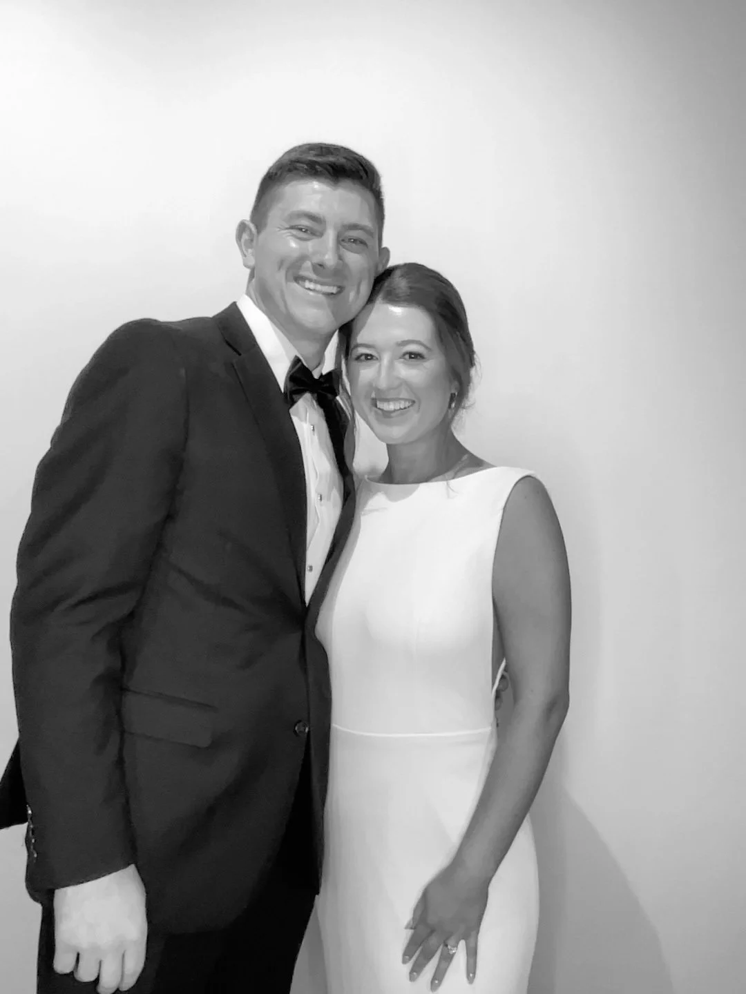A black and white photo of a bride and groom, posing in a photo booth. The groom is wearing a tuxedo with a bow tie, and the bride is in a sleeveless white dress, with her hair styled up.