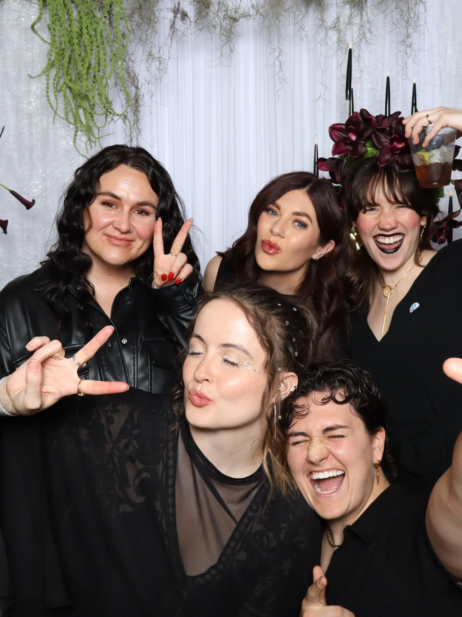 A group of women at a gala celebration, making peace signs and smiling in a photo booth with festive decorations and dark floral headpieces.