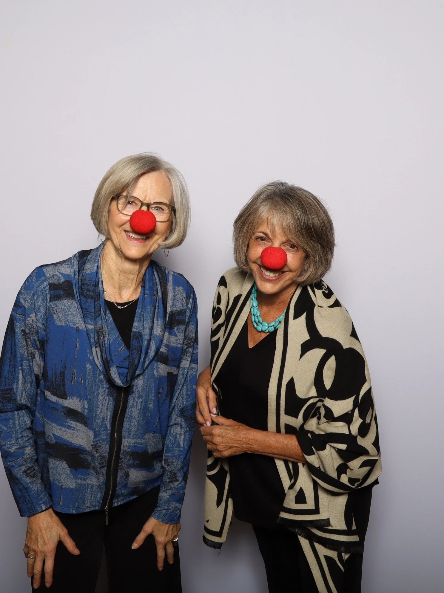 Two smiling women with red clown noses and casual attire standing against a plain white background in a photo booth.