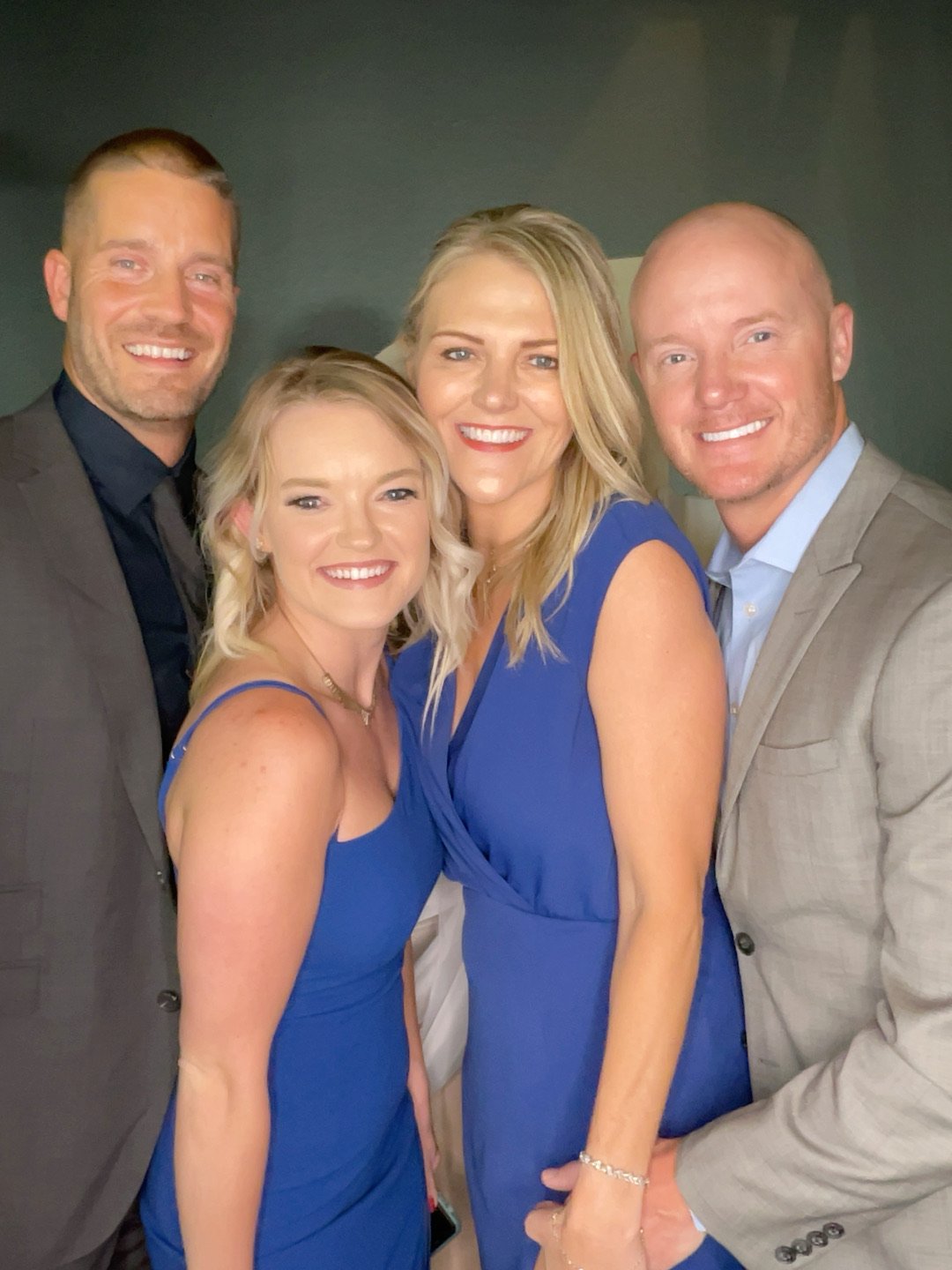 Group of five adults dressed in formal clothing smiling for a photo booth, two women in blue dresses and three men in suits, indoors against a dark background.