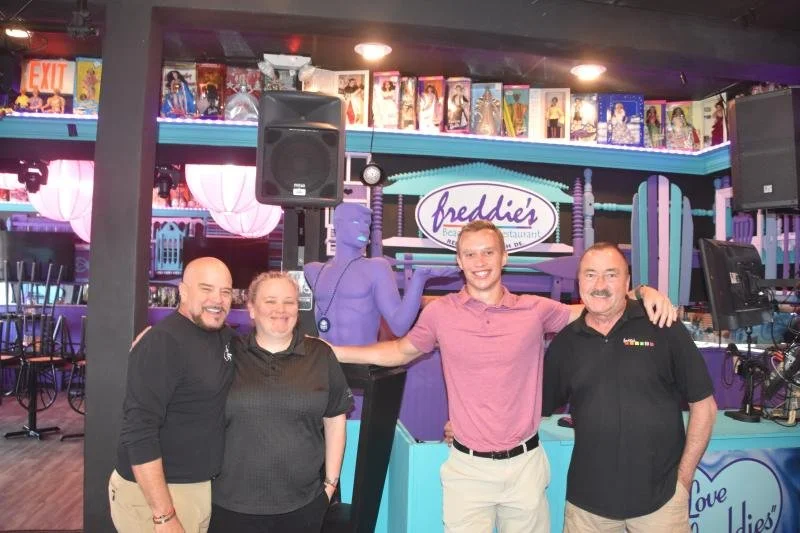 Four people standing together in a colorful arcade or entertainment venue named Freddie's, with a purple humanoid figure statue and arcade games in the background.