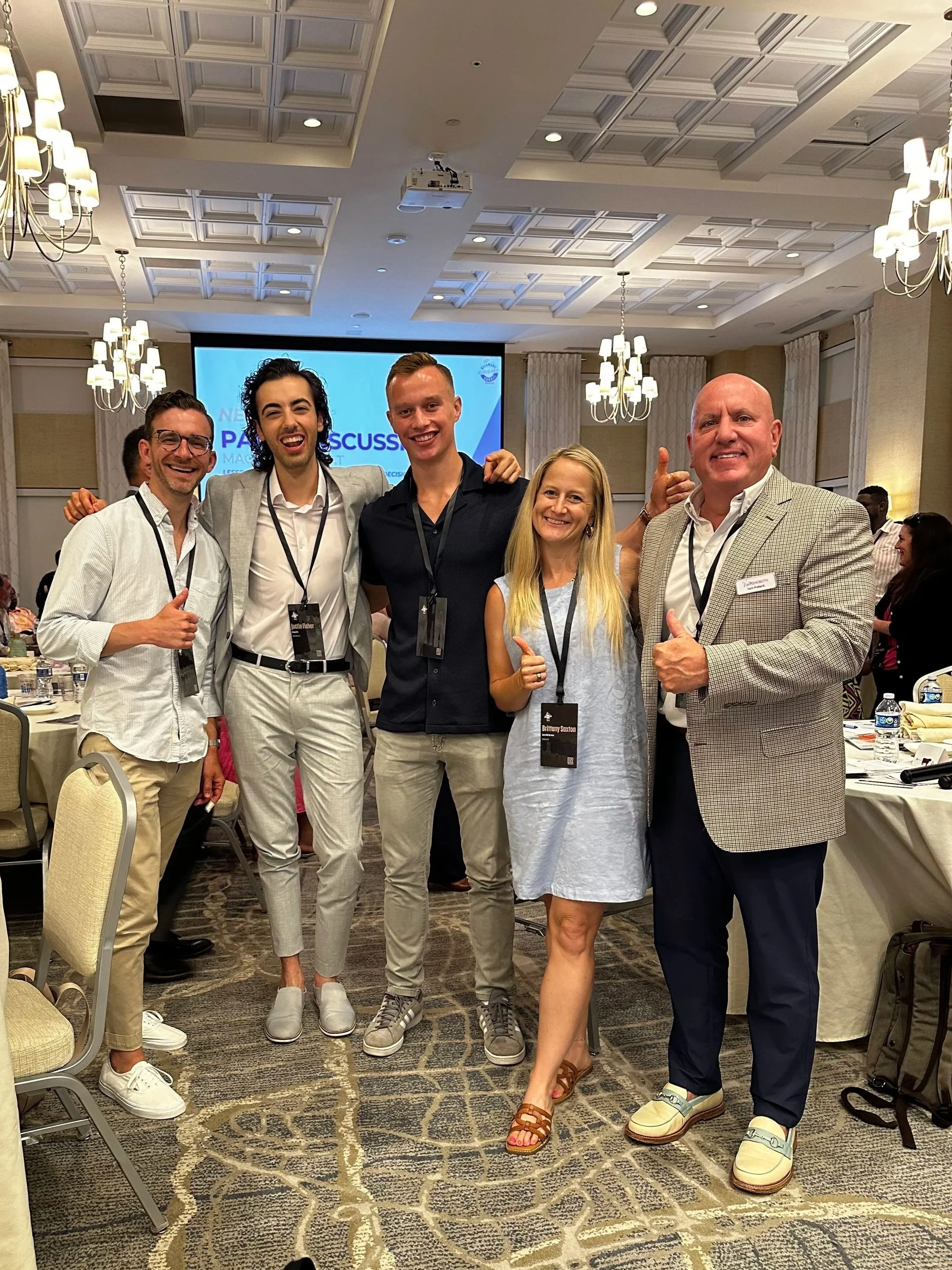 Group of five diverse people posing together at a conference, smiling and giving thumbs-up or pointing. The background shows a presentation screen and conference room setting.