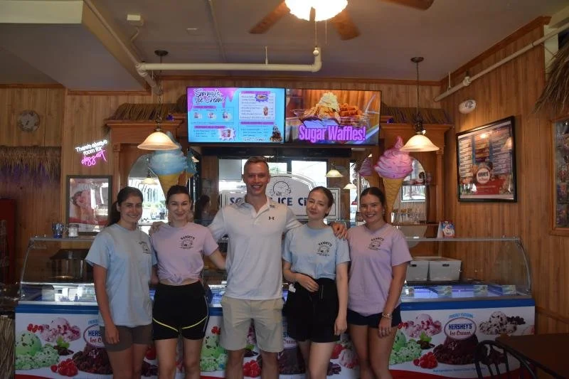 Group of five people posing inside ice cream shop, four young women and one young man, behind display freezers filled with ice cream, with menu screens and colorful decorations on wooden walls.