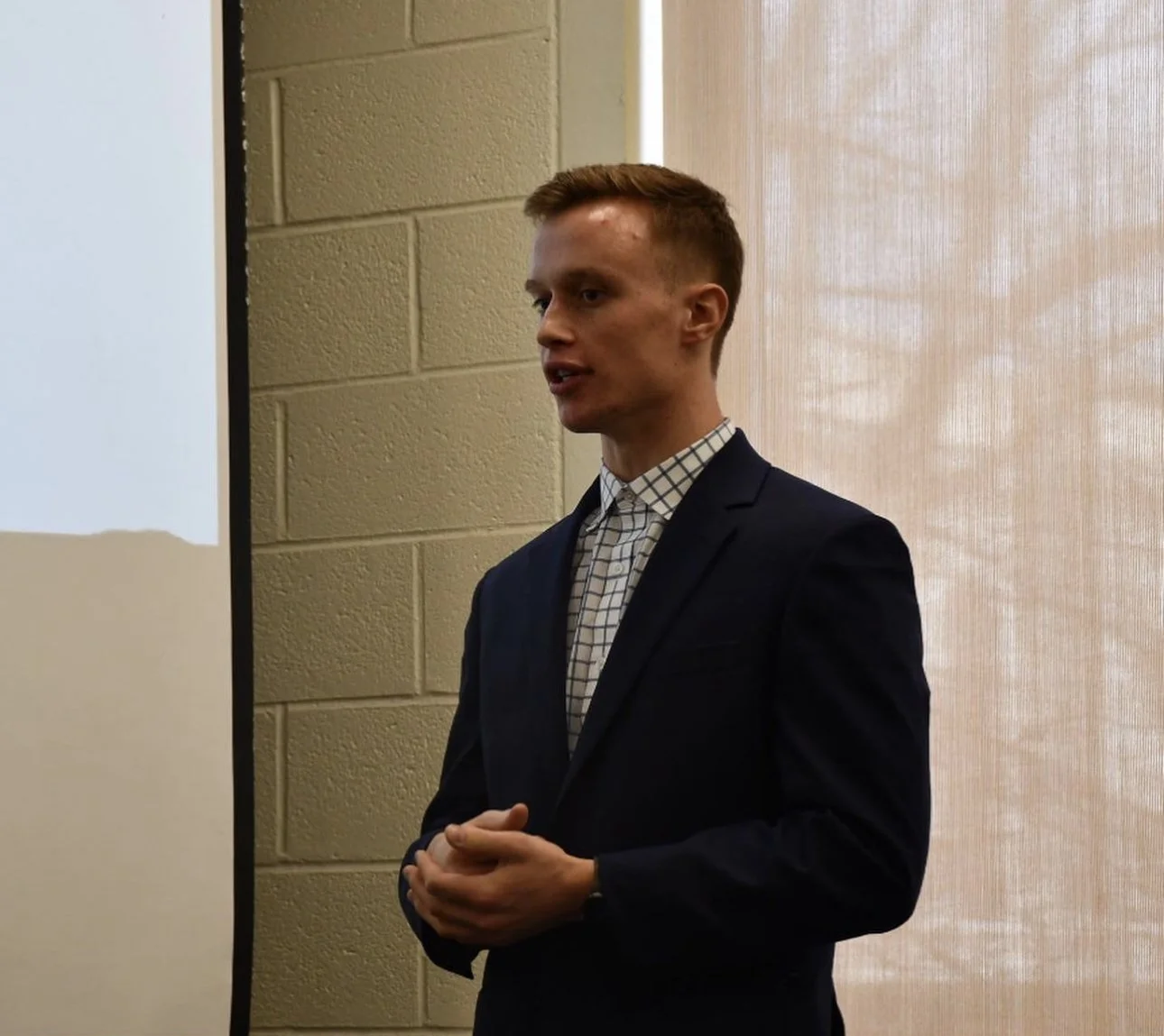 Young man in a dark suit and checkered shirt giving a presentation in a classroom or conference room.