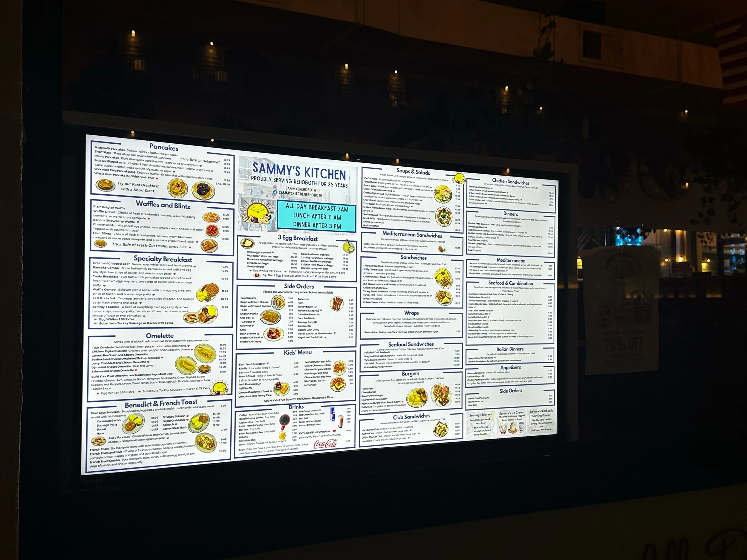 illuminated restaurant menu board displaying breakfast, lunch, dinner, sandwiches, soups, salads, and other offerings at Sammy's Kitchen with some yellow smiley face icons and a small Coca-Cola advertisement at the bottom.