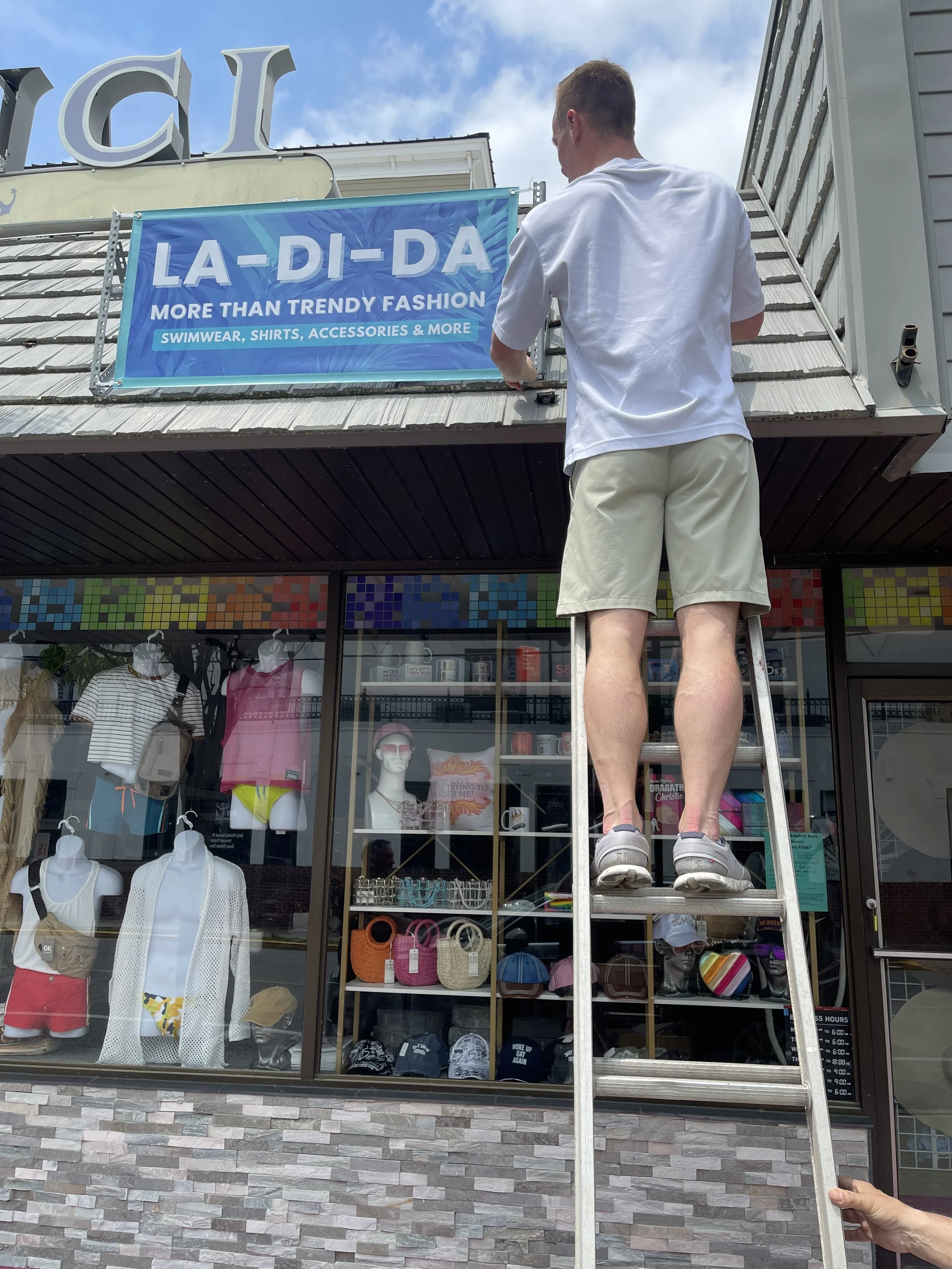 A man standing on a ladder putting up a large blue banner that reads 'LA-DI-DA MORE THAN TRENDY FASHION SWIMWEAR, SHIRTS, ACCESSORIES & MORE' outside a retail store.