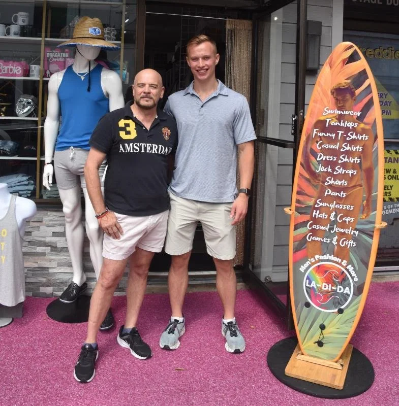 Two men standing in front of a surfboard-shaped sign outside a store, with a mannequin wearing a blue tank top and gray shorts behind them. The sign lists summer accessories and clothing items such as swimsuits, tank tops, and hats.