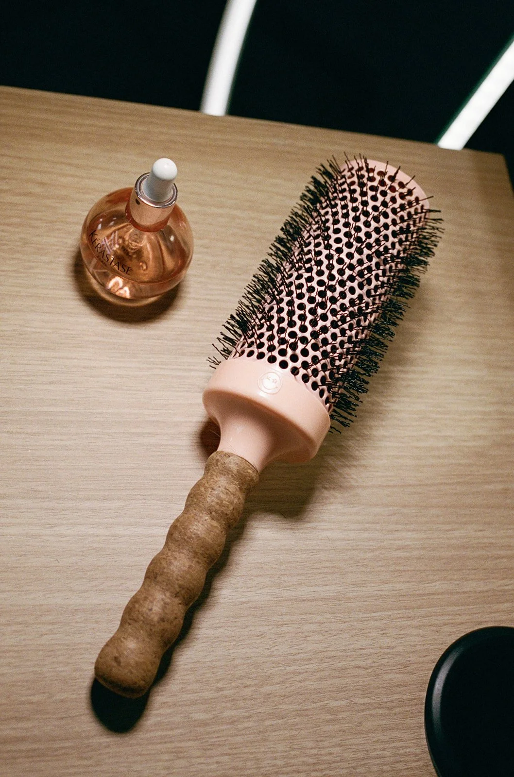 A round hairbrush with a light pink handle and black bristles, a small bottle of pink hair oil, and a black container on a wooden surface.
