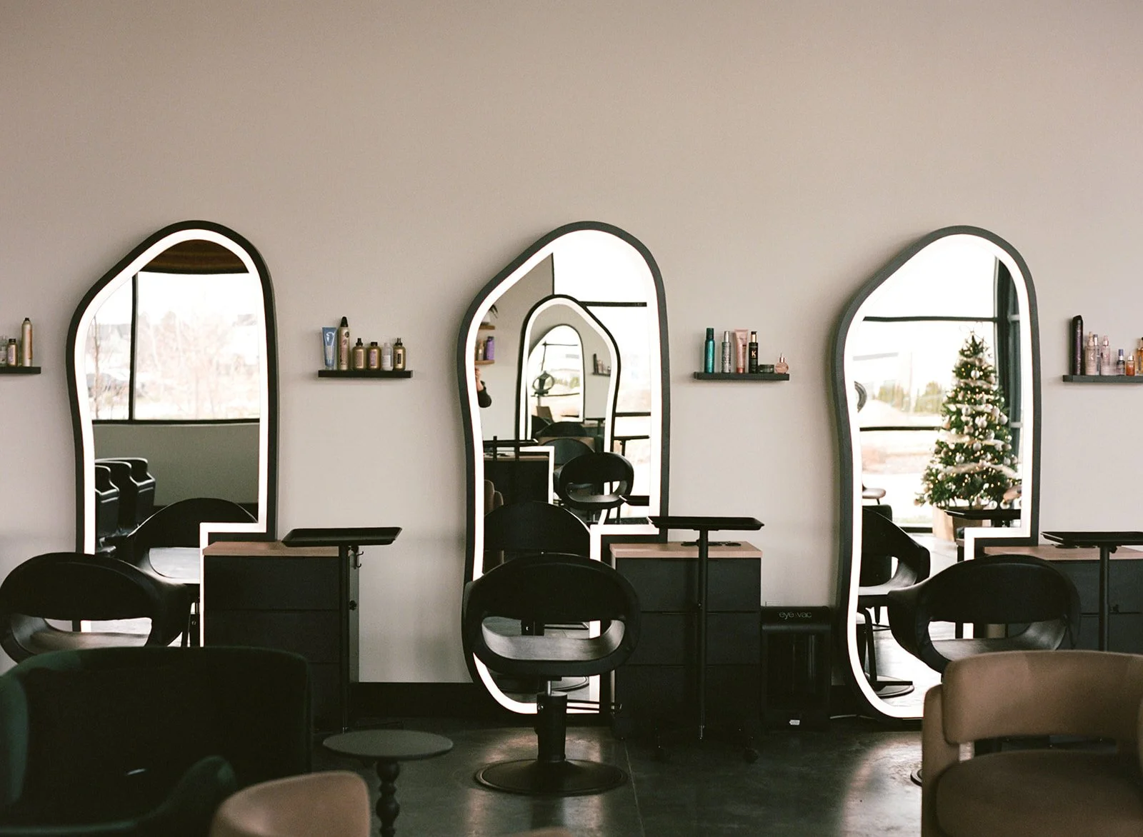 Interior of a hair salon with three stylists' stations, each with a black chair, mirror, and shelves of hair products, decorated with a Christmas tree visible through the window.