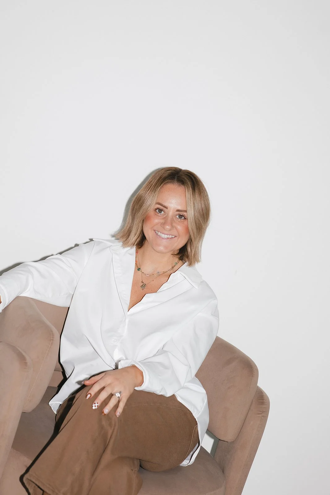 A woman with shoulder-length blonde hair, smiling, sitting on a beige armchair against a white background, wearing a white button-up shirt and brown pants.