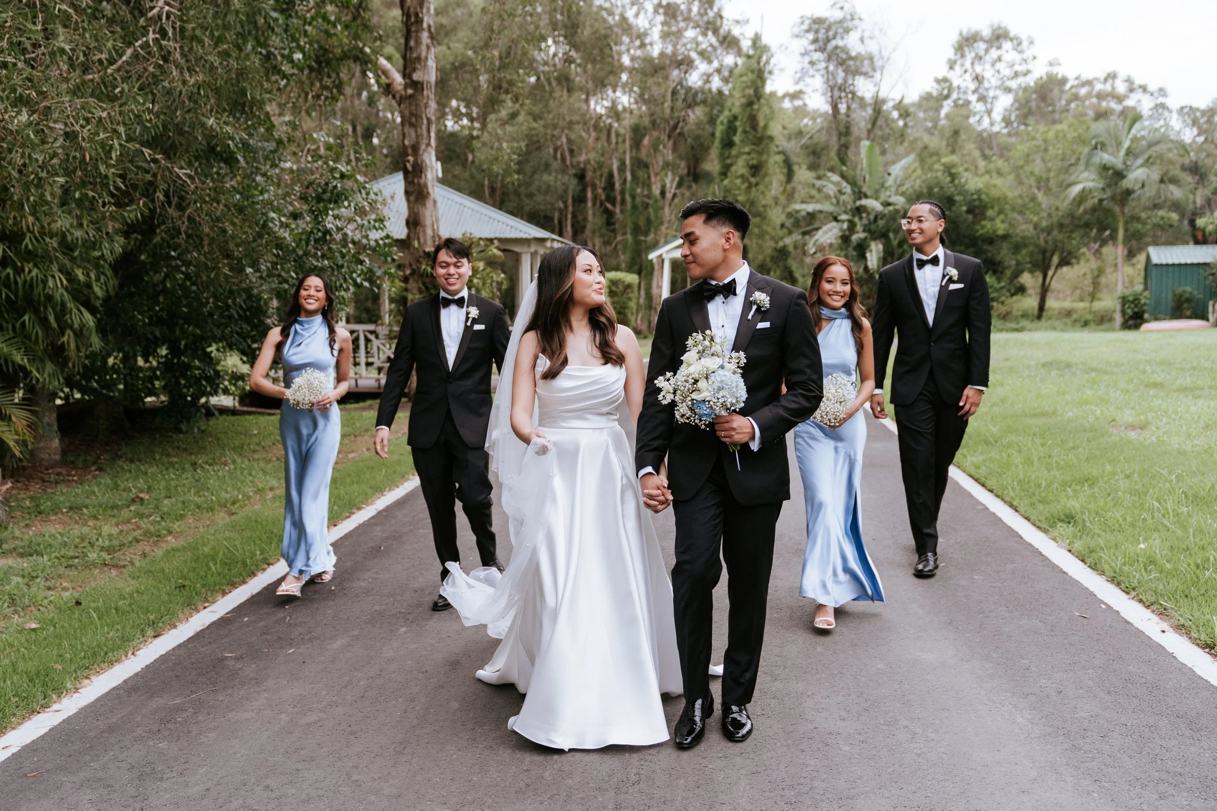 Bride and groom walking with bridesmaids in blue dresses and groomsmen in tuxedos outdoors on a path, surrounded by trees.