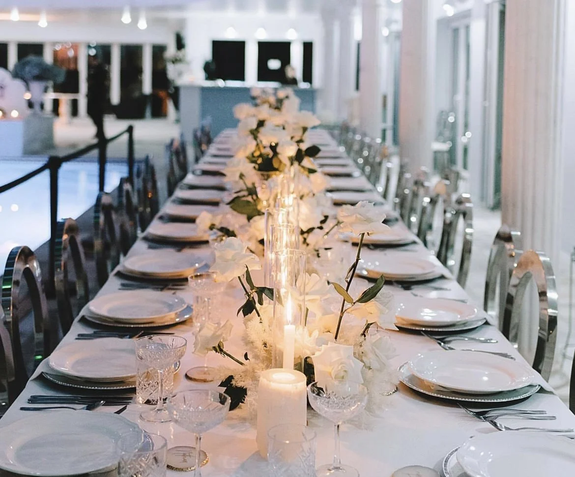 Elegant wedding table setting with white tablecloth, plates, crystal glassware, candles, and floral centerpieces featuring white roses.