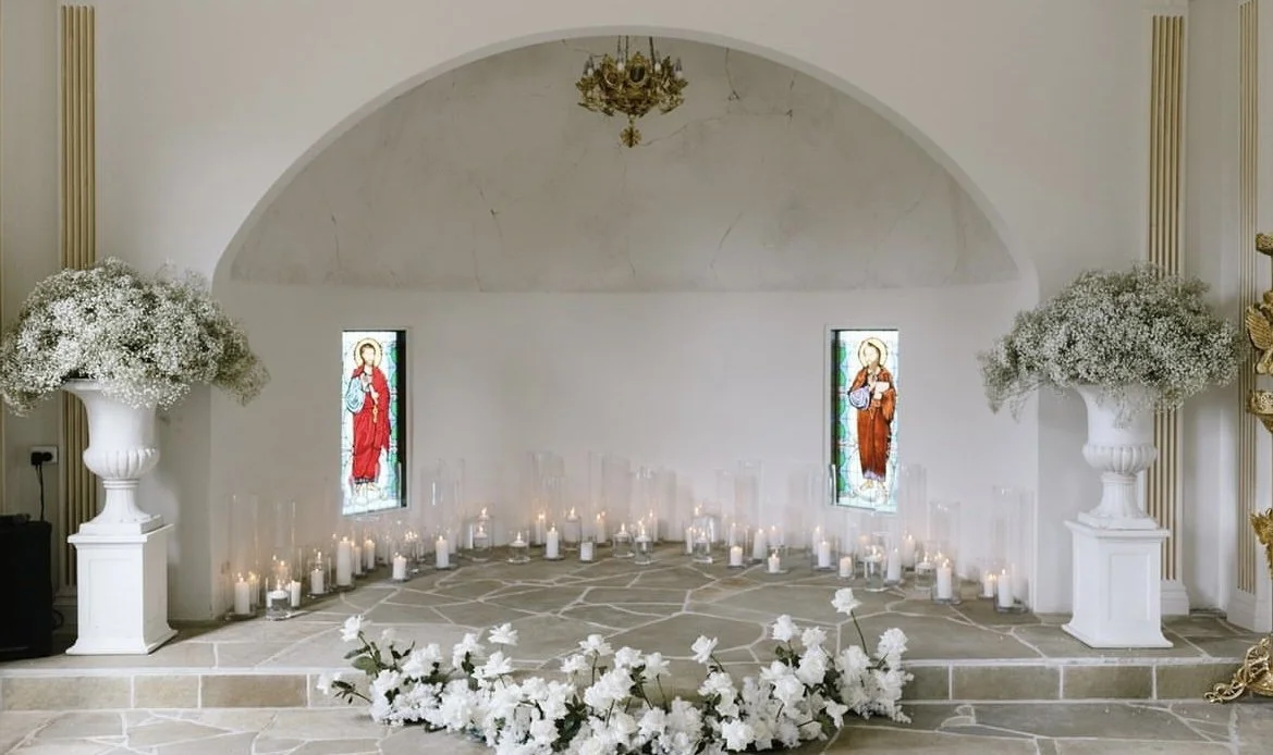 Interior of a chapel with stained glass windows, lit candles, large floral arrangements, and a stone platform.