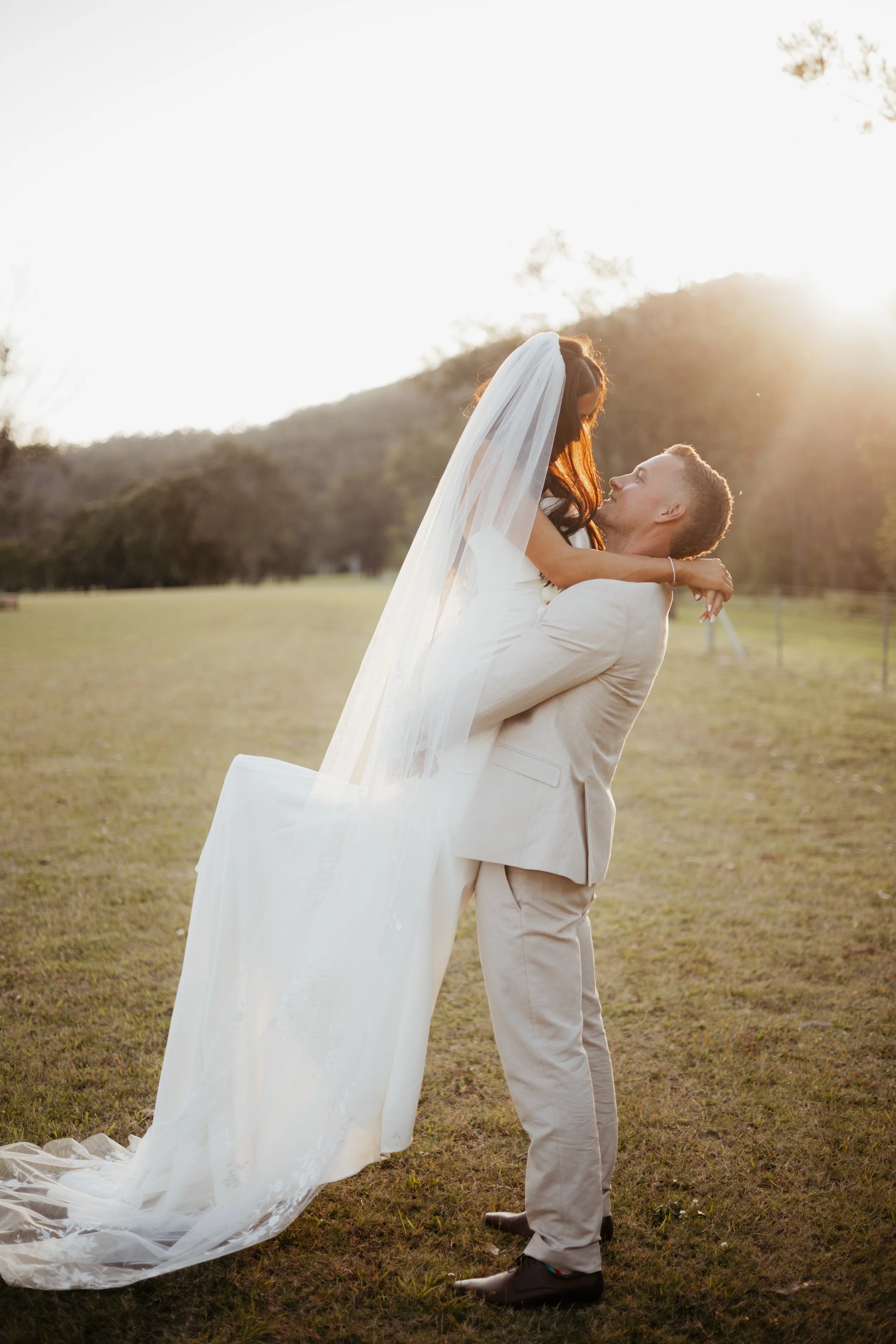 A groom lifting and embracing a bride in a wedding dress with a long veil in an outdoor setting during sunset.